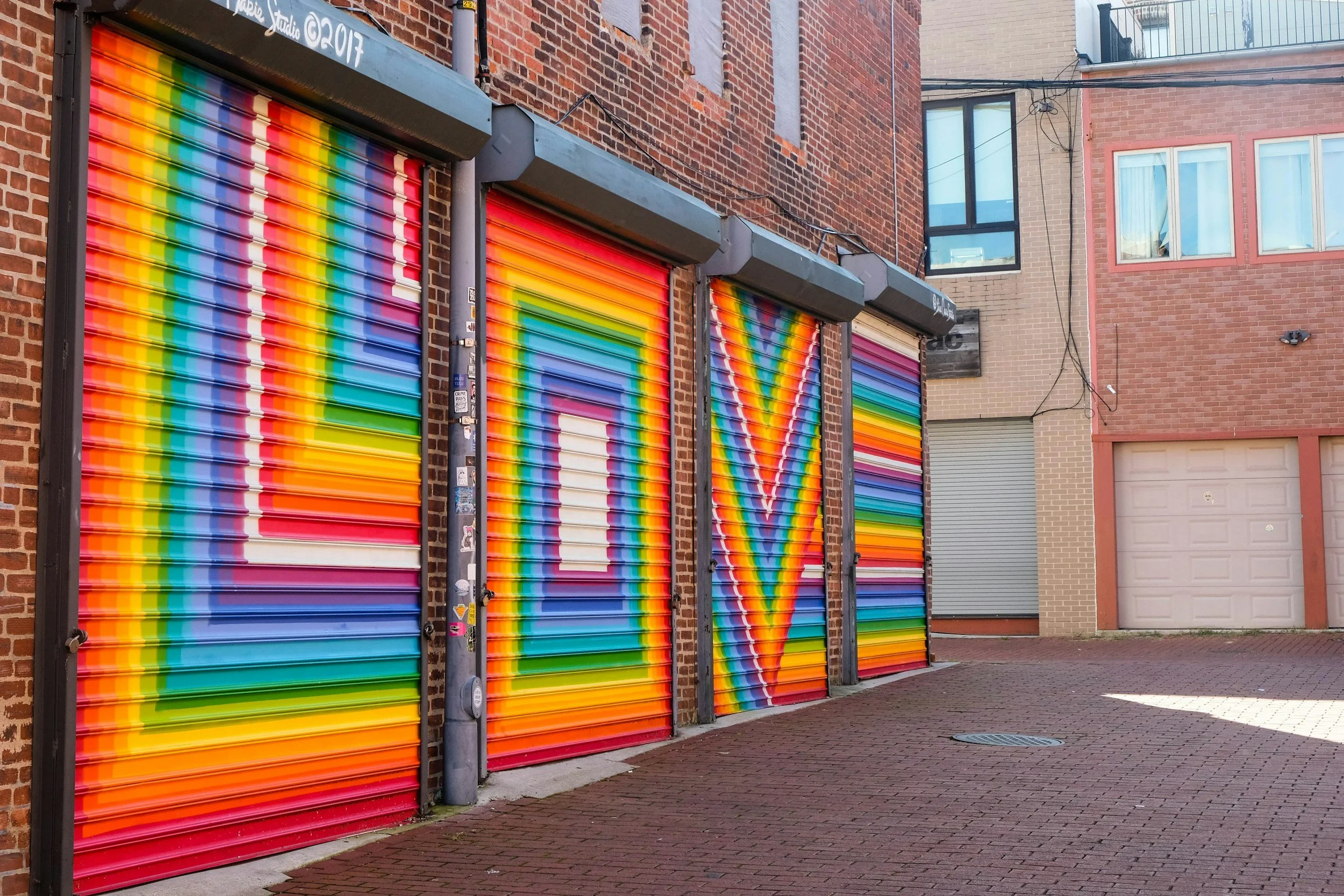 Four colorful rainbow- striped metal shutters on a brick building with a brick-paved alleyway in front.