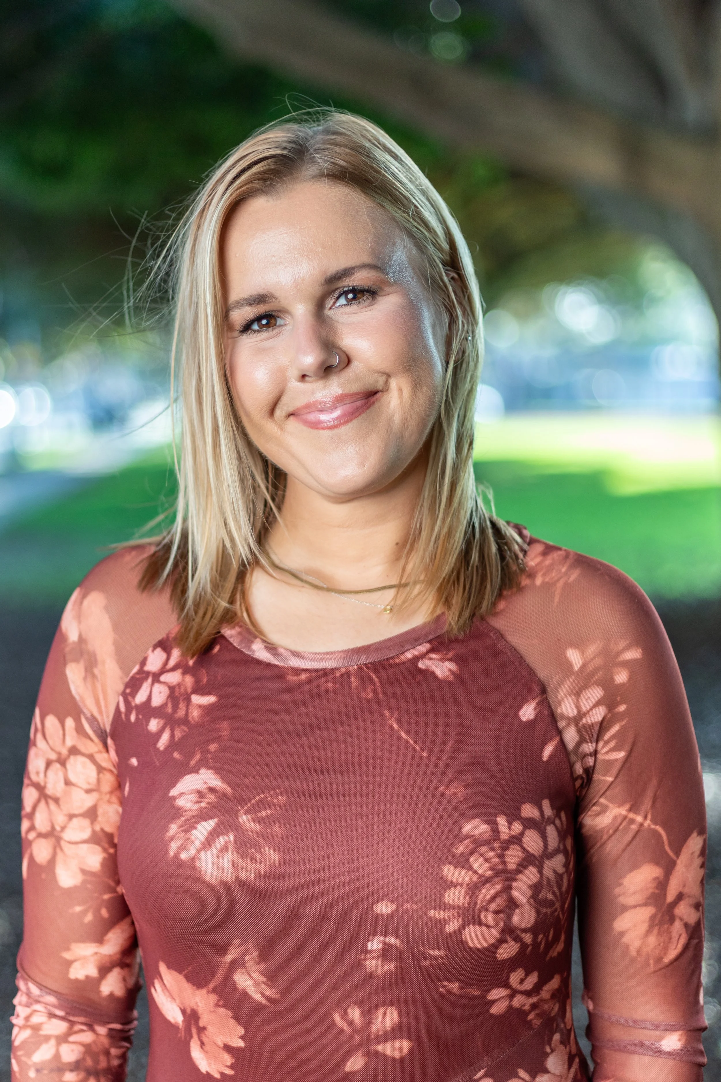 A smiling young woman with blonde hair and a nose ring, standing outdoors with a blurred background of green grass and trees.