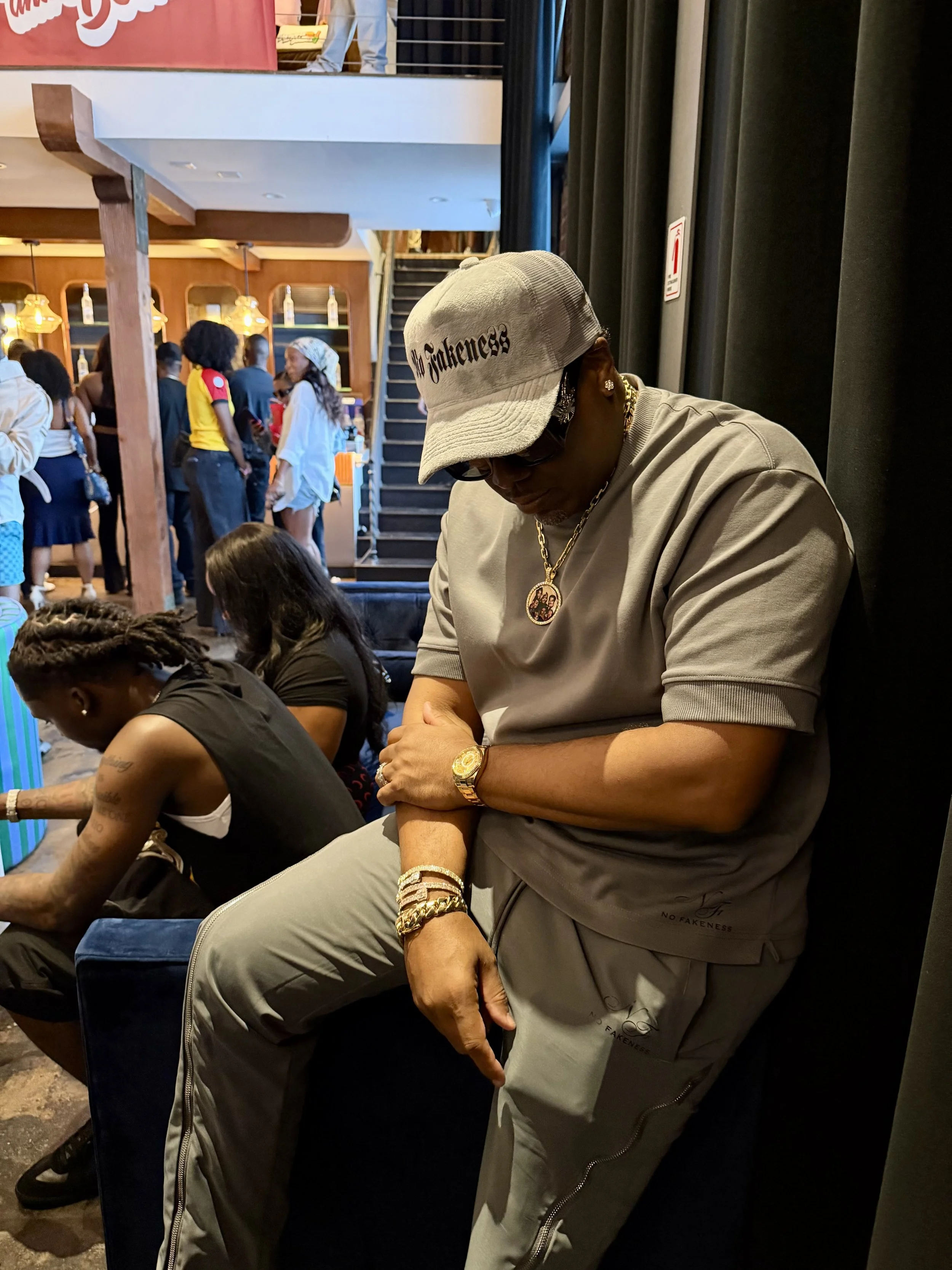 A man sitting indoors with his head lowered, wearing a gray cap, sunglasses, gold jewelry, and gray clothing. In the background, there are people standing and a staircase leading upwards.