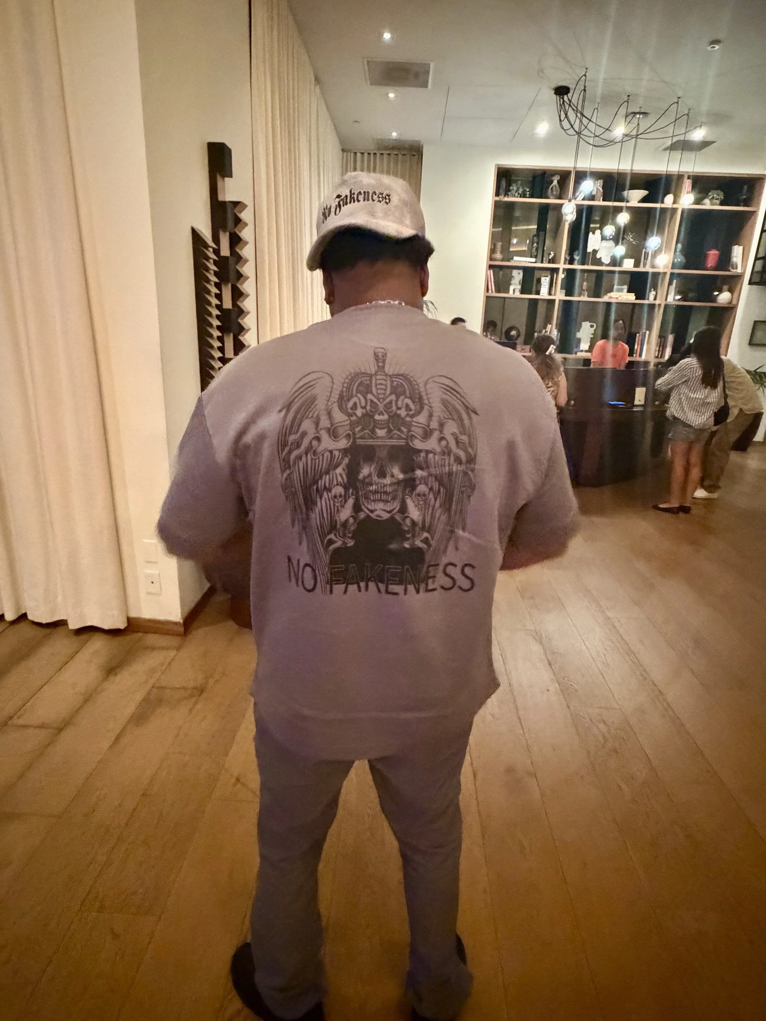 Back view of a person wearing a white graphic T-shirt with a skull design and the phrase "NO FEARLESS" and a white baseball cap with the words "no fakeness" in black, standing in a dimly lit room with wooden flooring and a bookshelf in the background.