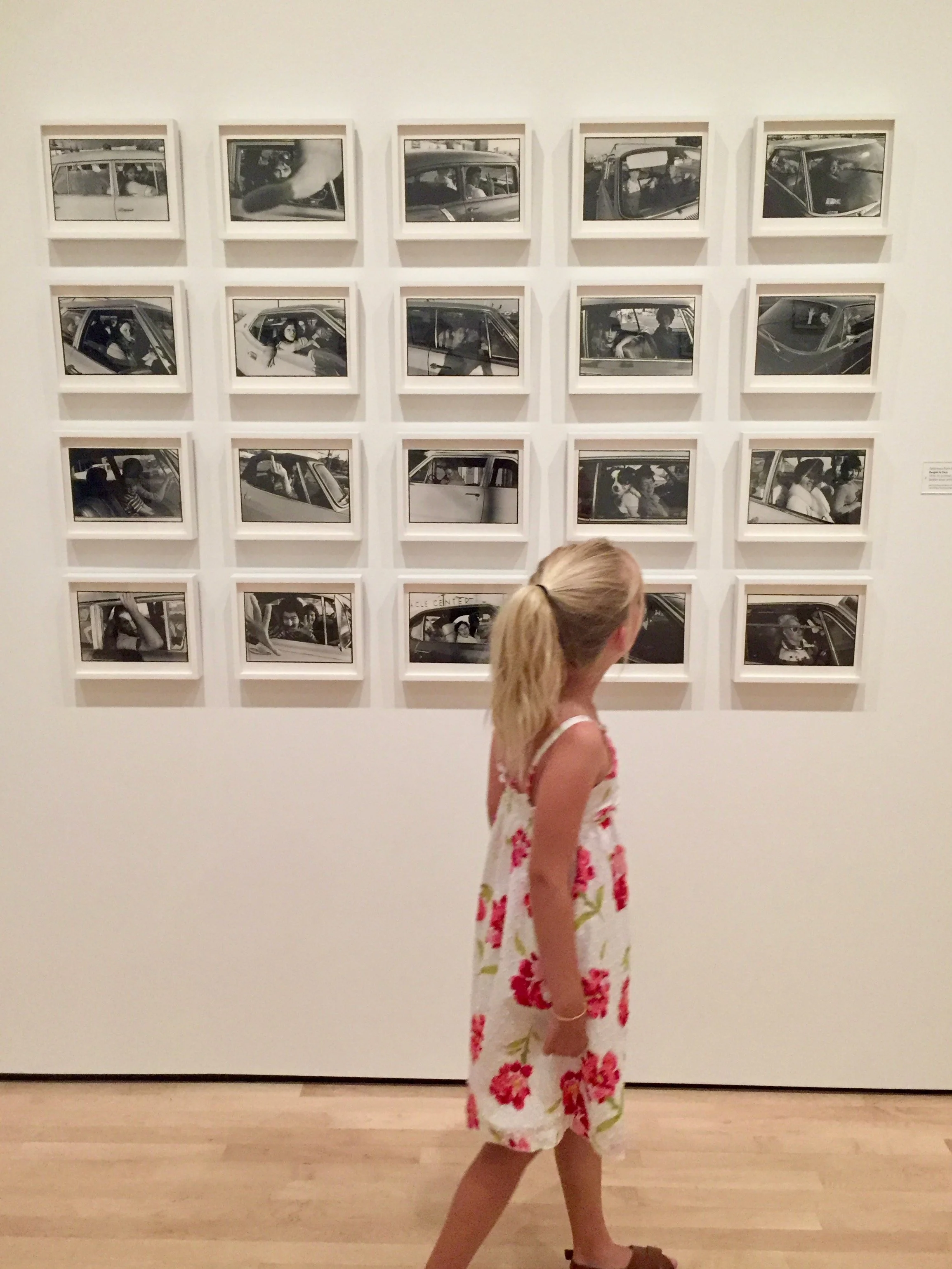 A young girl walking in front of a wall display of black and white photographs in frames.