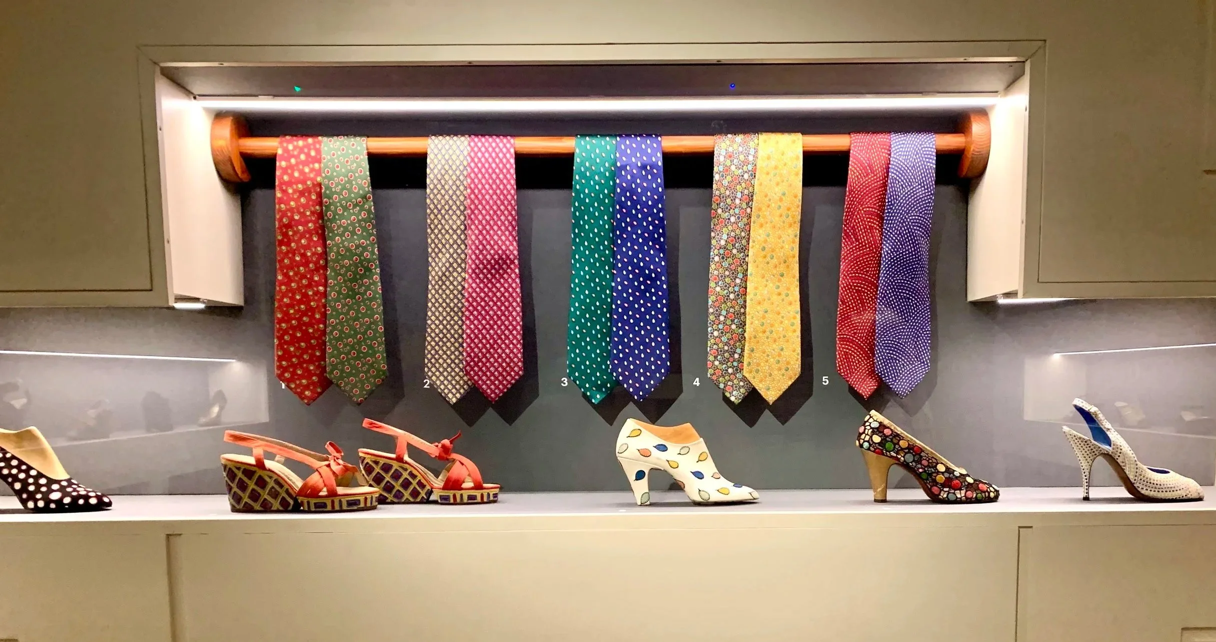 Display of six colorful patterned neckties hanging on a rod and six decorated high-heeled shoes on a shelf below, with a gray background.