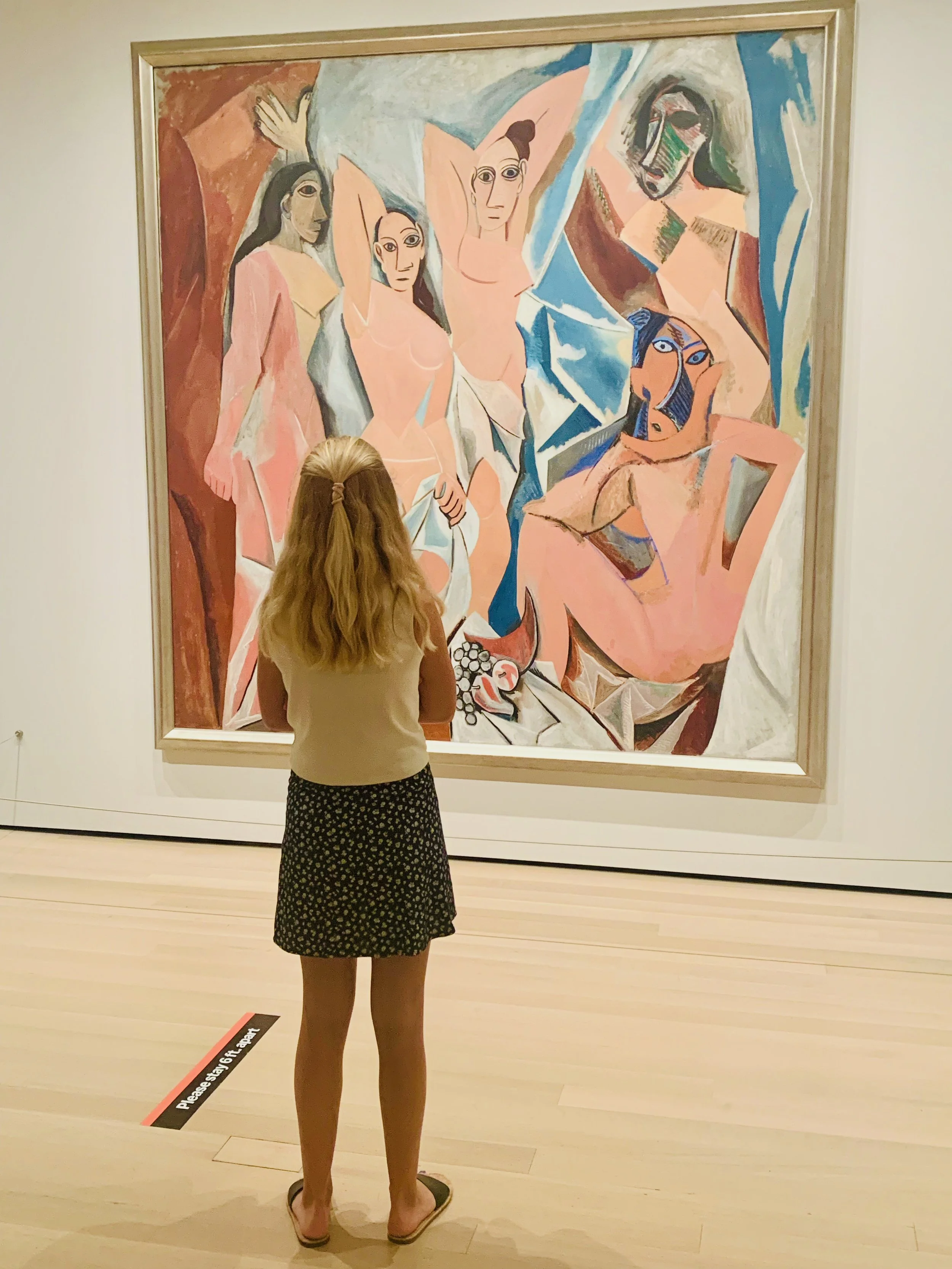 A girl standing in front of a large abstract painting by Pablo Picasso of five women with varied skin tones and hair styles, in MOMA New York.