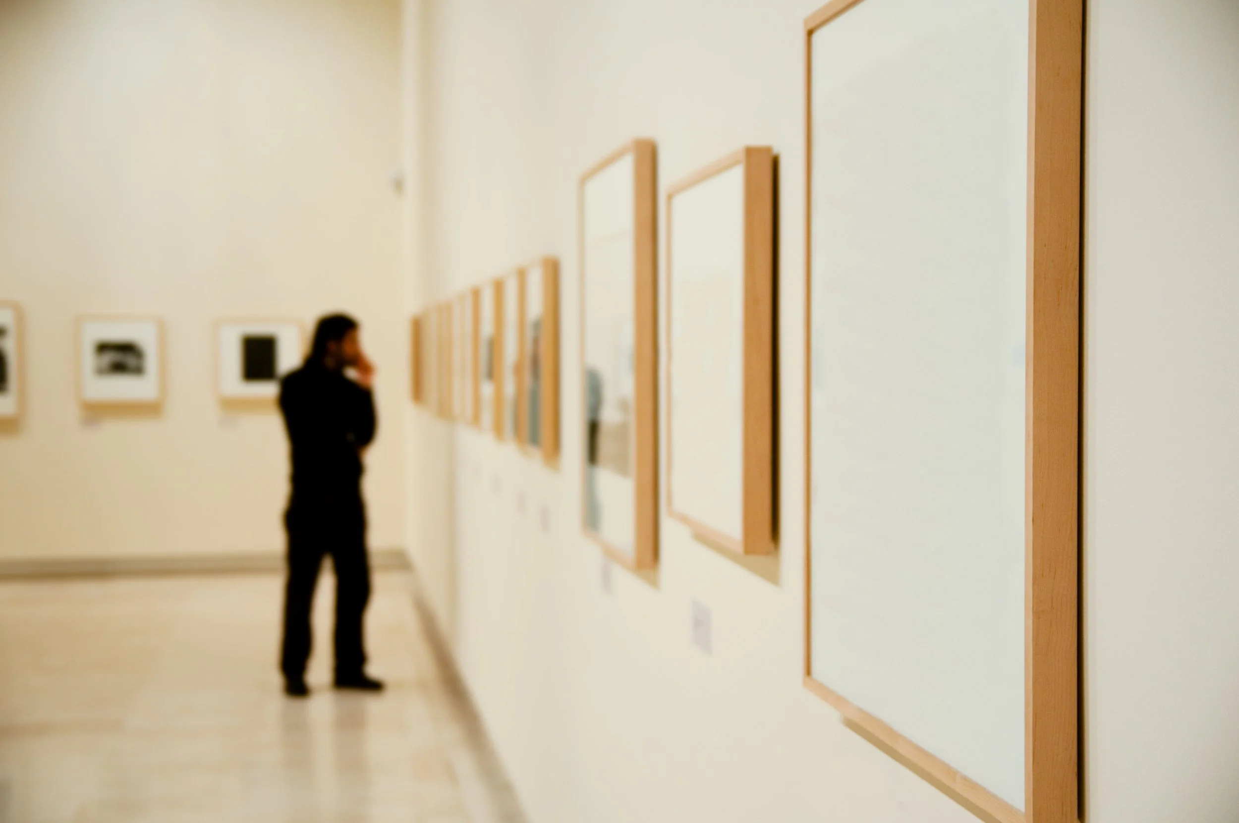 A person in a black outfit looking at framed artwork on a white gallery wall.