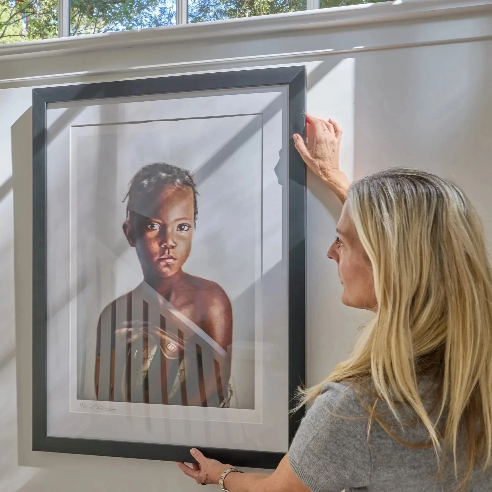 Arts consultant Laura Bradley hangs a framed portrait of a young black girl with solemn expression on a white wall inside a bright room with a window.