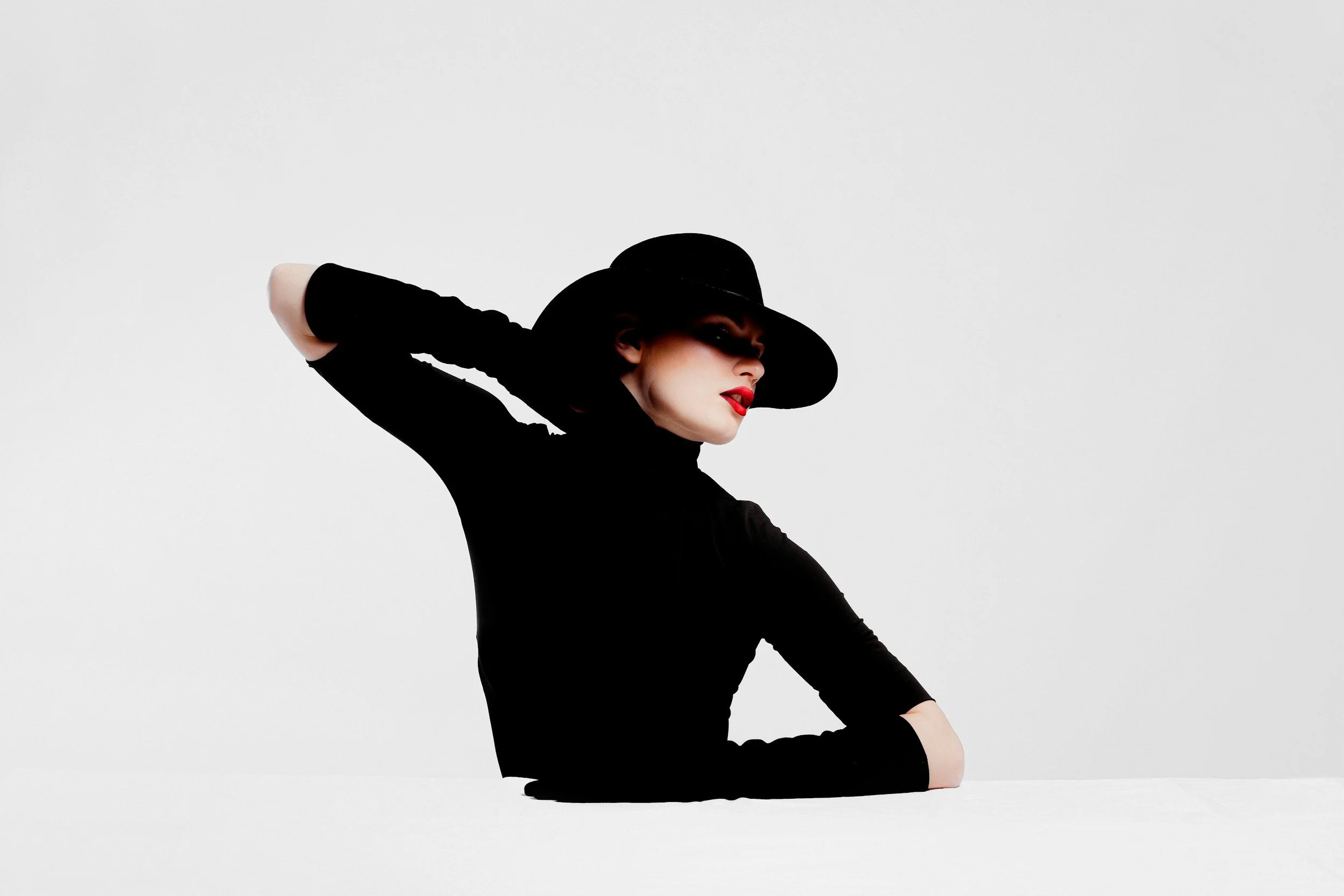 A woman dressed in black with red lipstick, wearing a wide-brimmed hat, resting her head and arm on her hand, against a plain light background.