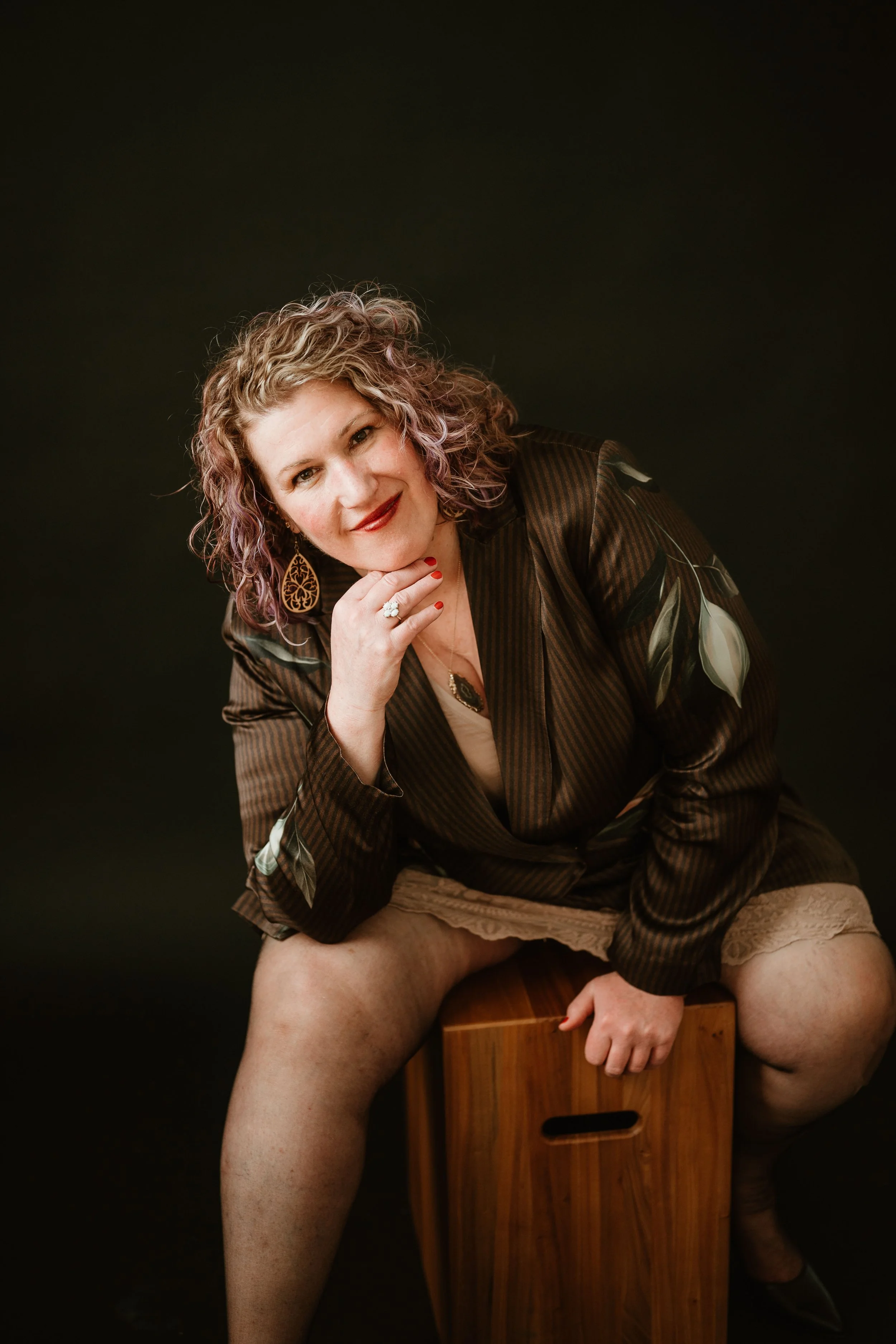 A woman with curly blonde hair and earrings is sitting on a wooden box, smiling at the camera with her hand on her chin. She is wearing a brown striped blazer with floral patterns, red lipstick, and has red nail polish. Her legs are exposed and she is wearing beige lace shorts and black shoes. The background is black.