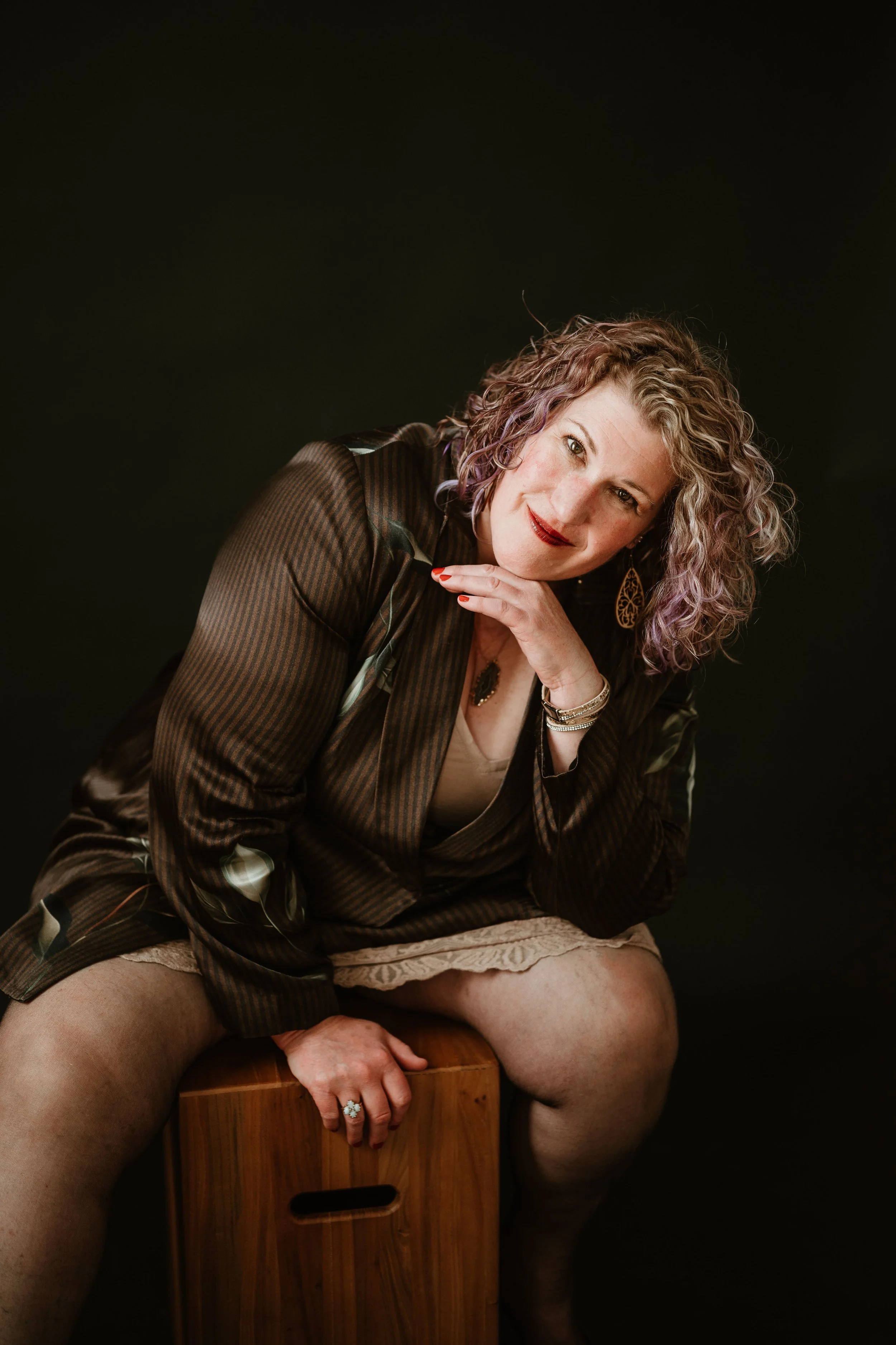 A woman with curly hair, wearing a striped blazer and beige shorts, sitting on a wooden box, smiling and resting her chin on her hand, against a black background.