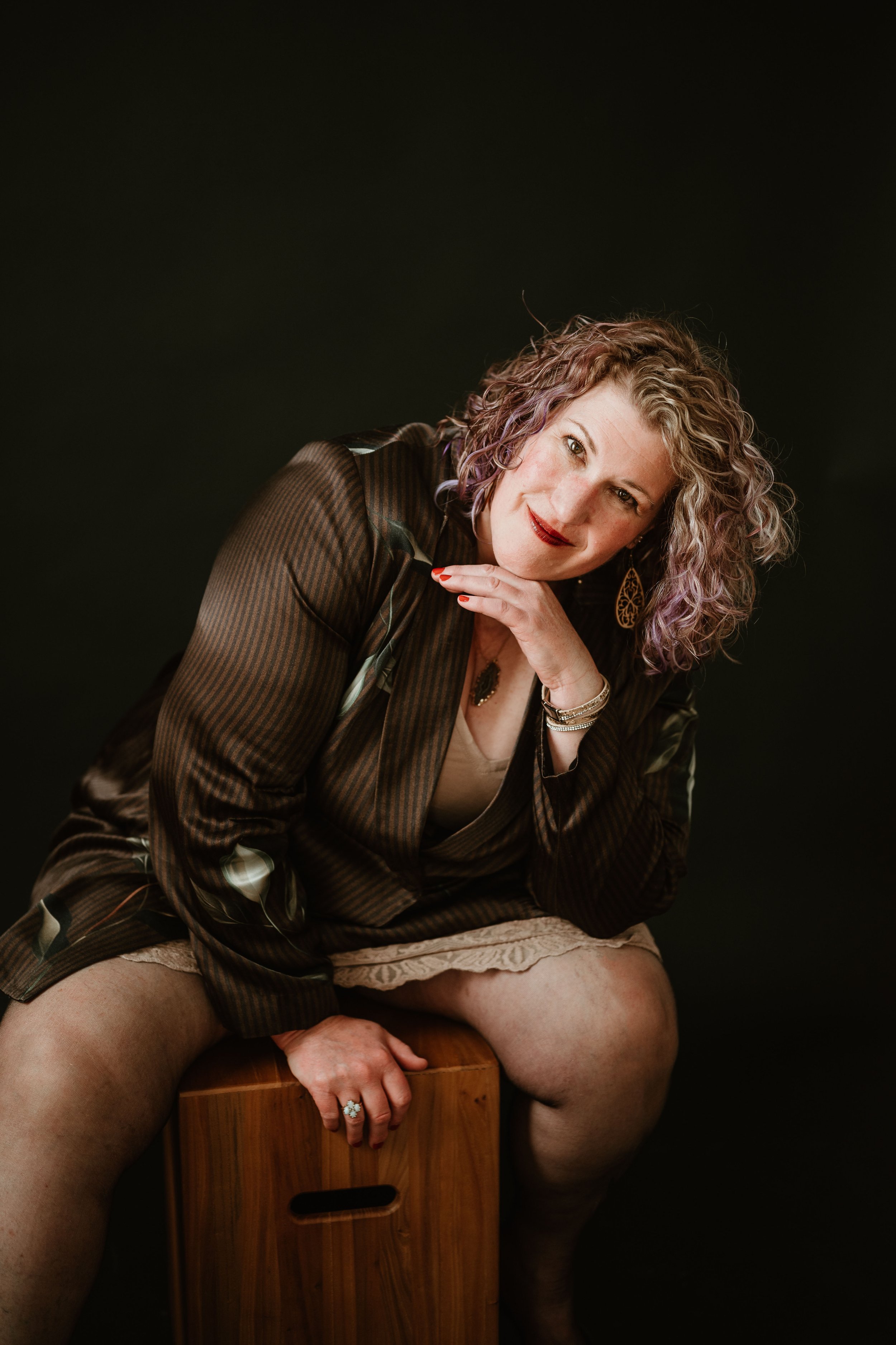 A woman with curly hair and red lipstick sitting on a wooden box against a black background. She is wearing a striped jacket, beige lace shorts, and various jewelry, and is posing with her head resting on her hand.