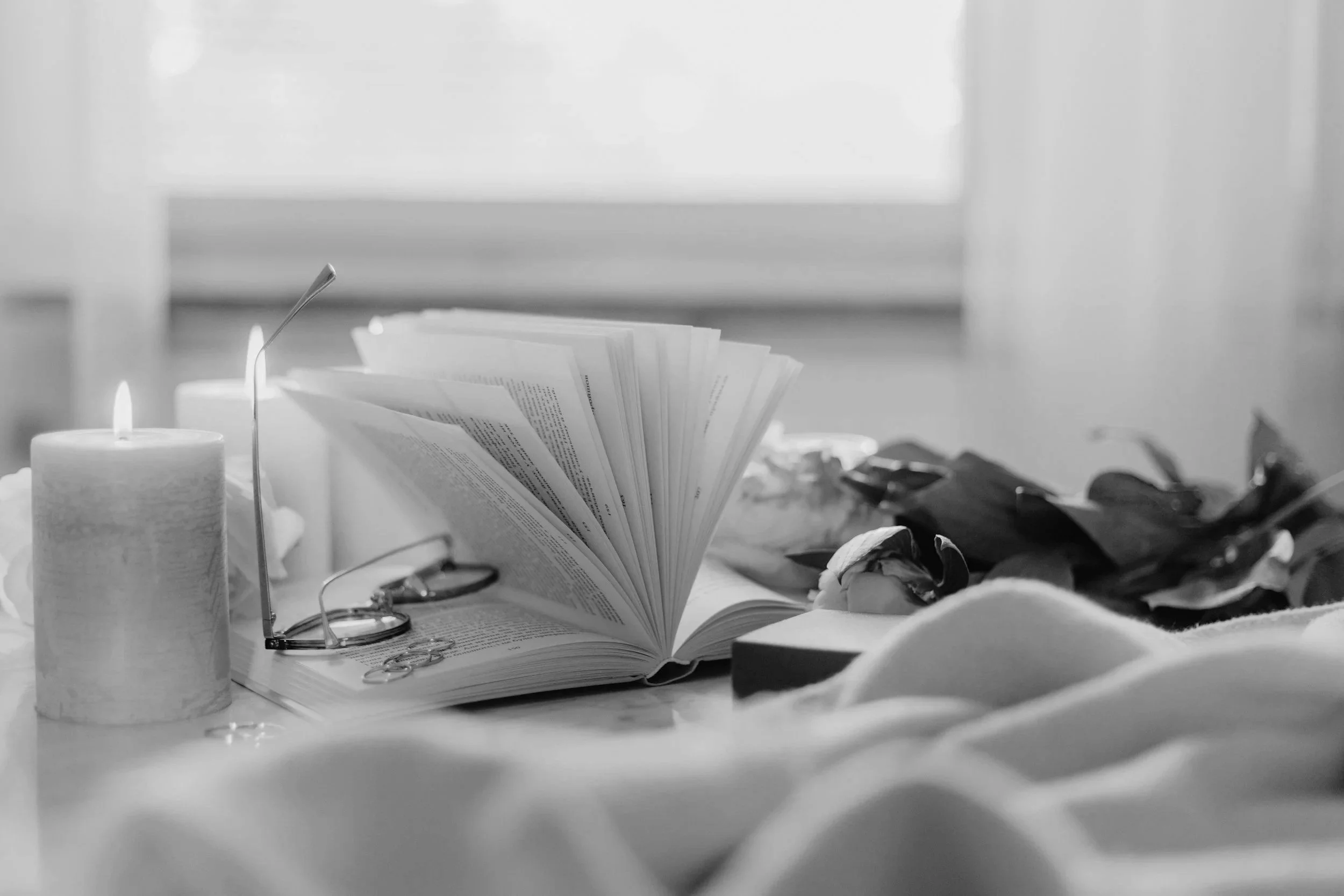 A black and white photo of an open book with reading glasses resting on it, candles, scissors, and scattered papers on a table, with a window in the background.