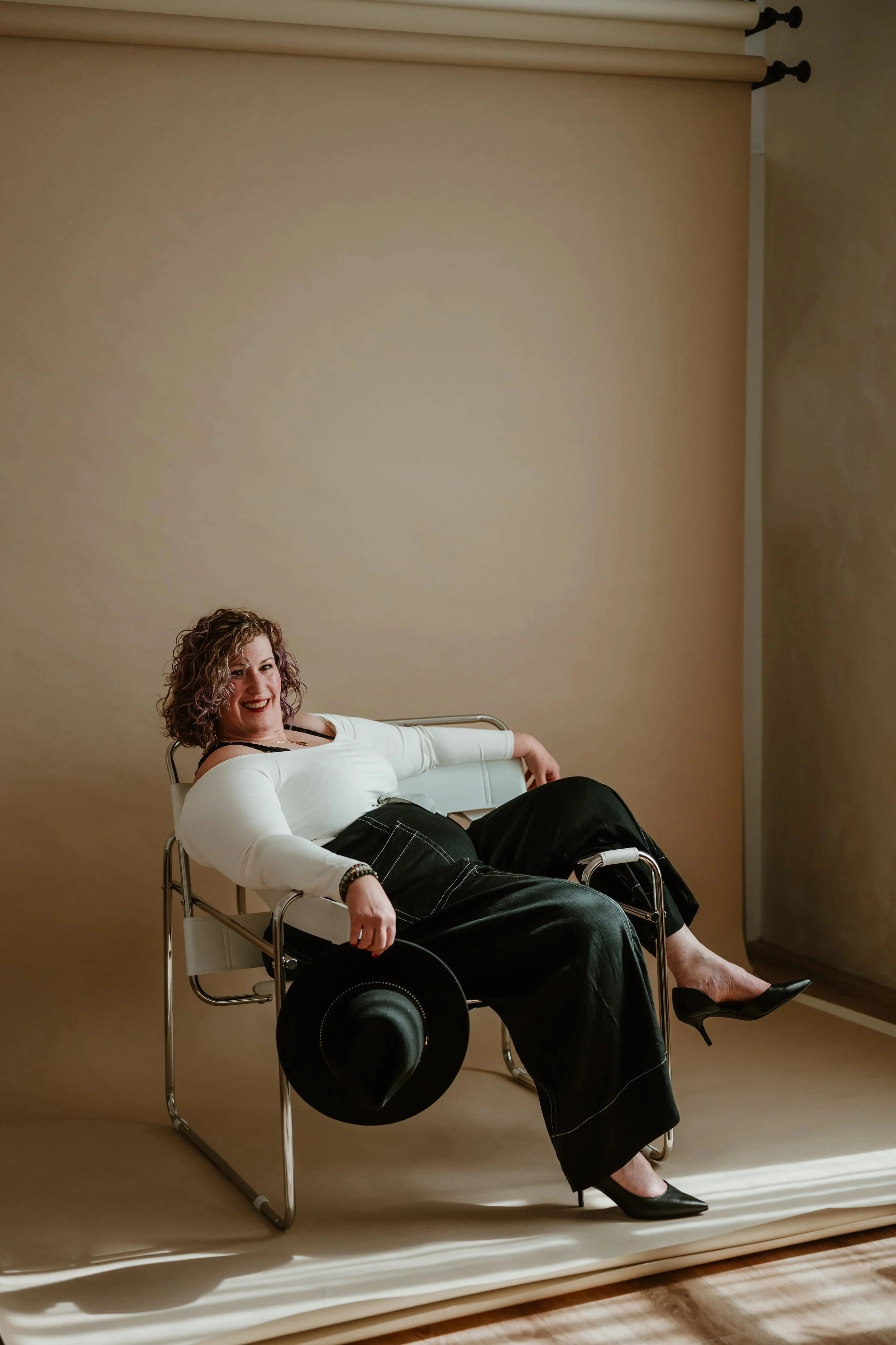A woman with curly hair, wearing a white top and black pants, sitting in a wheelchair with her legs crossed, smiling, against a beige wall background.