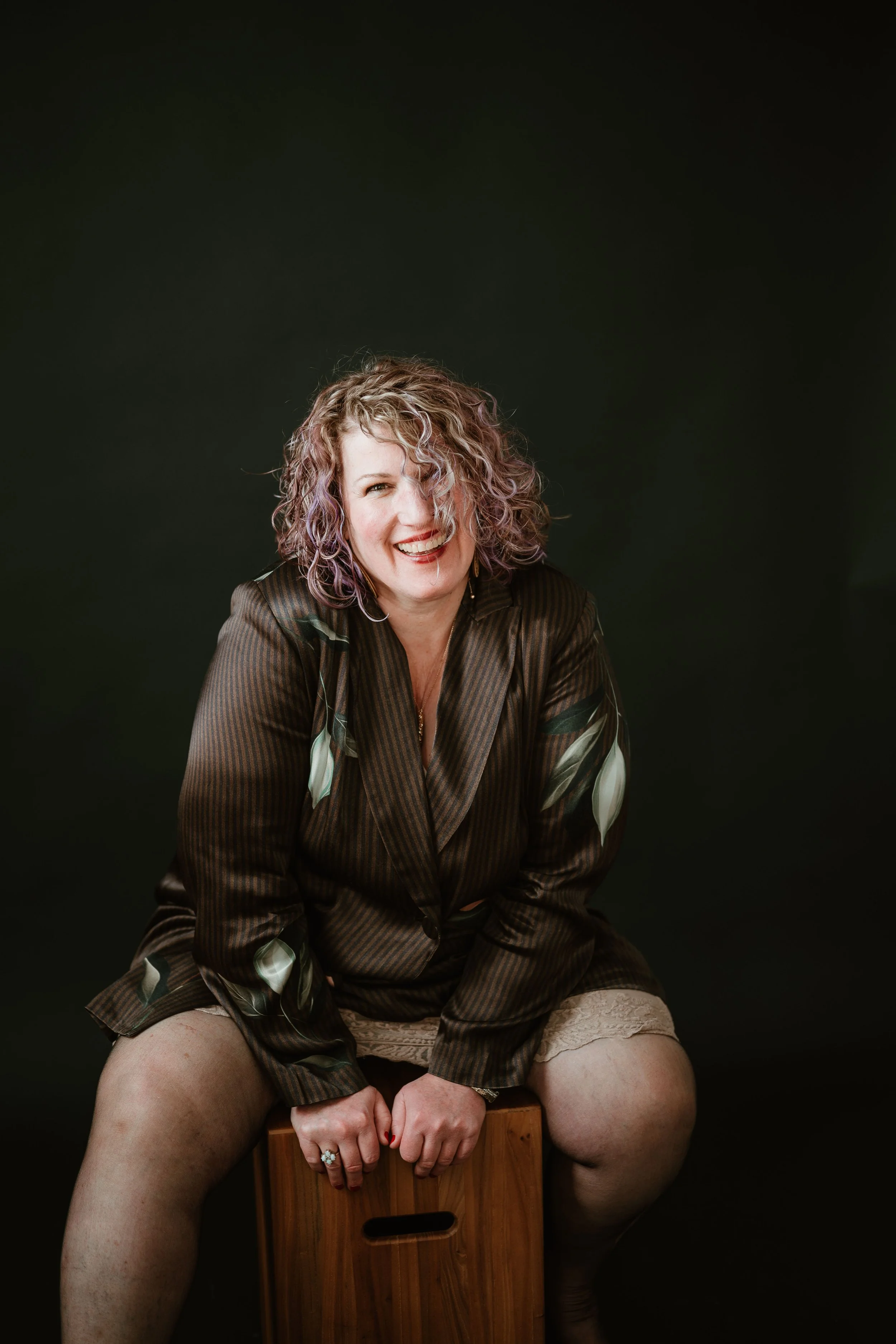 A woman with curly, purple-tinted hair sitting on a wooden stool, smiling and wearing a silk striped robe with floral patterns, against a black background.