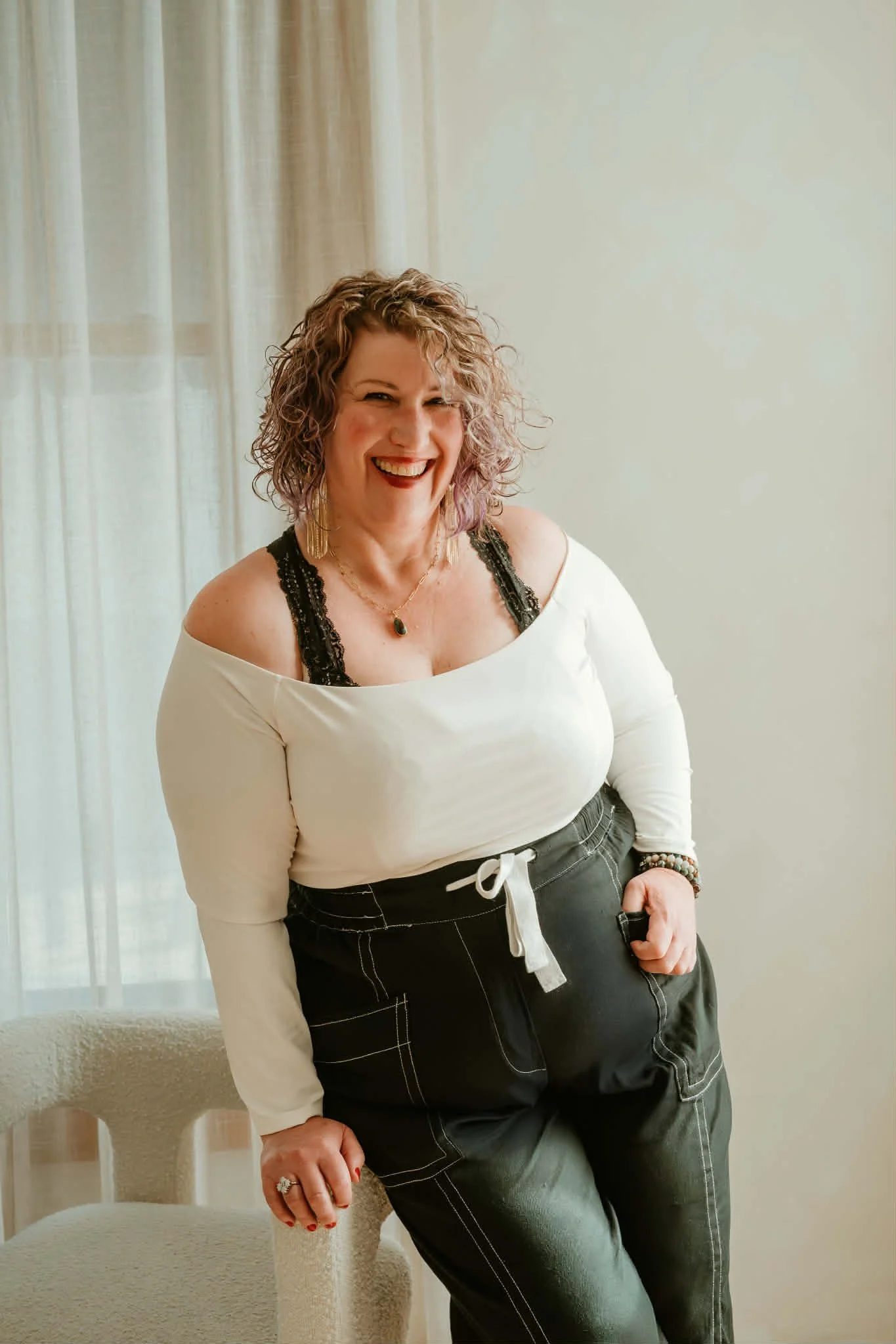 A woman with curly hair smiling and standing indoors near a chair, wearing a white off-shoulder top with black lace straps, black pants with white stitching, and jewelry.