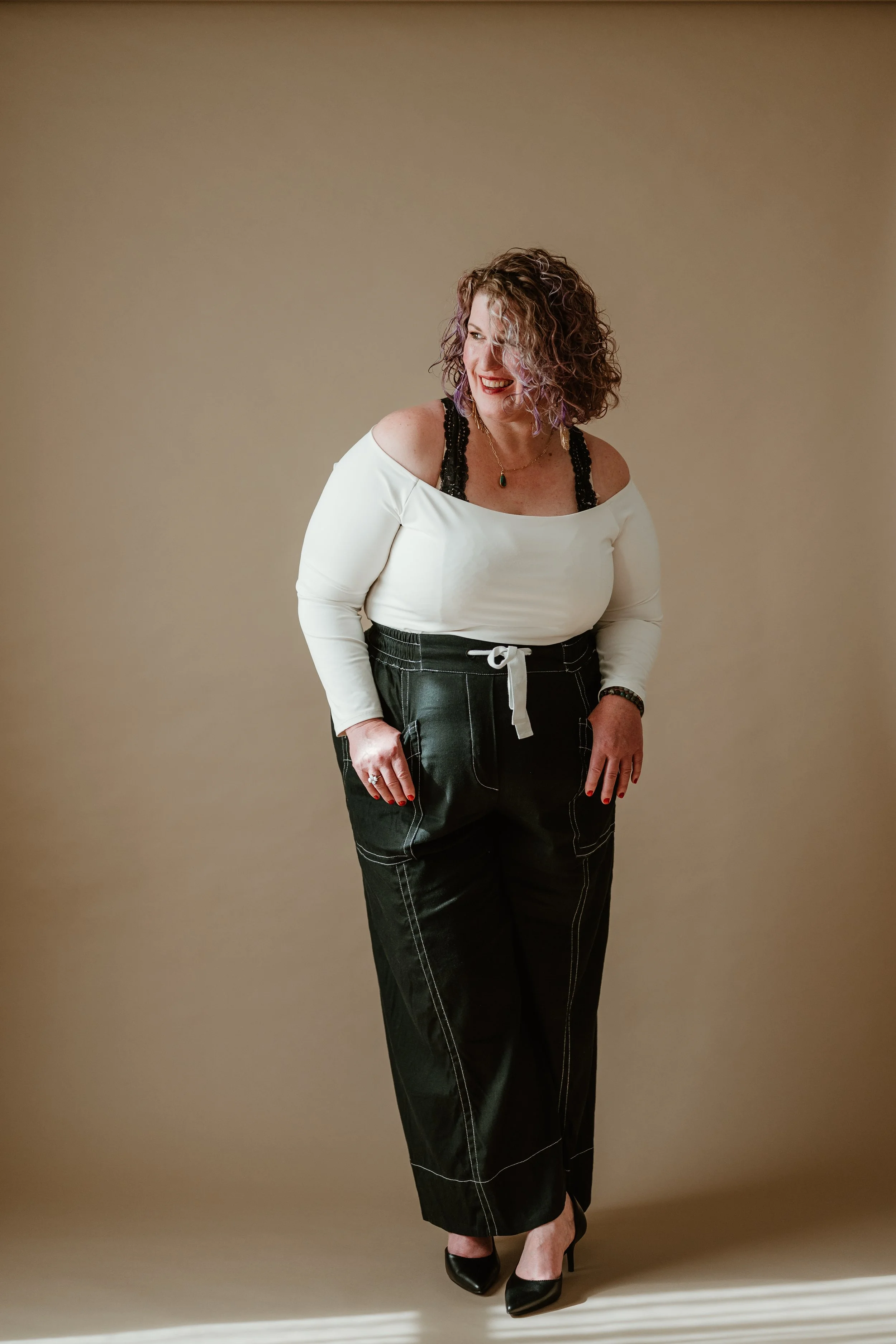 A woman with curly hair, wearing a white off-shoulder top with black lace straps, black high-waisted pants with white stitching, black high heels, and a green pendant necklace, standing against a beige background, smiling and looking to her right.