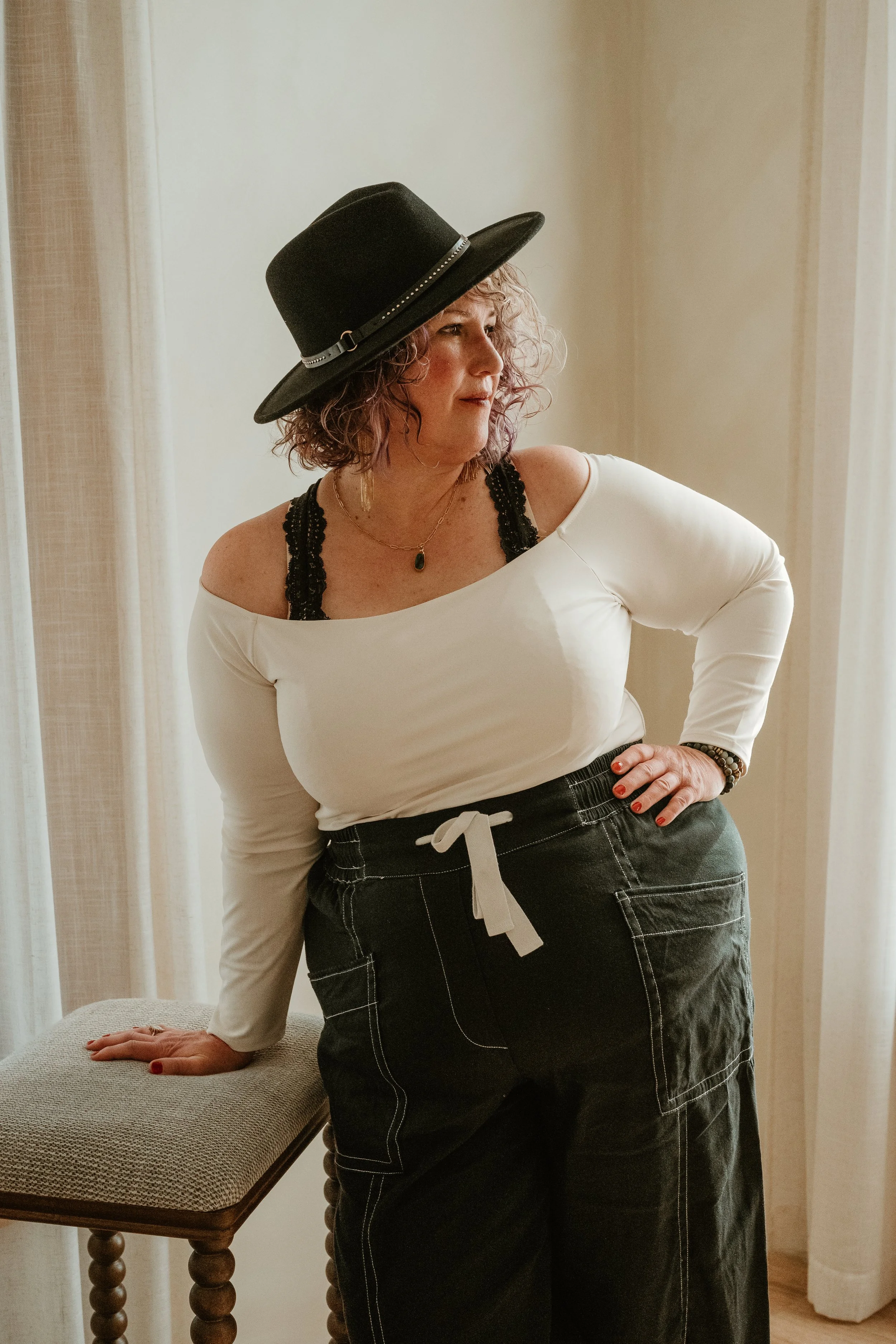 A woman with curly hair wearing a wide-brimmed black hat, an off-shoulder white top with lace straps, and black pants with white stitching standing indoors by a window and a bench.