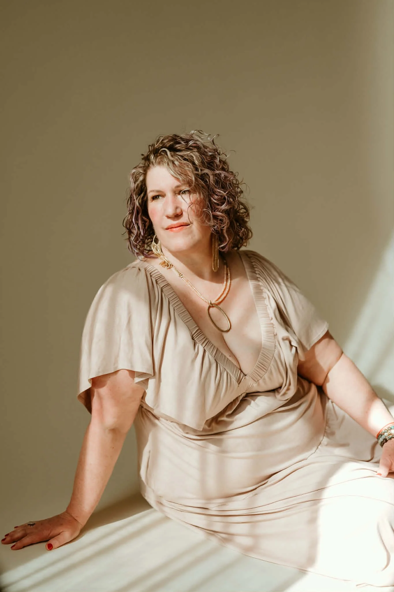 Page with curly brown hair sitting on the floor, wearing a mauve dress and jewelry, looking slightly to the side.