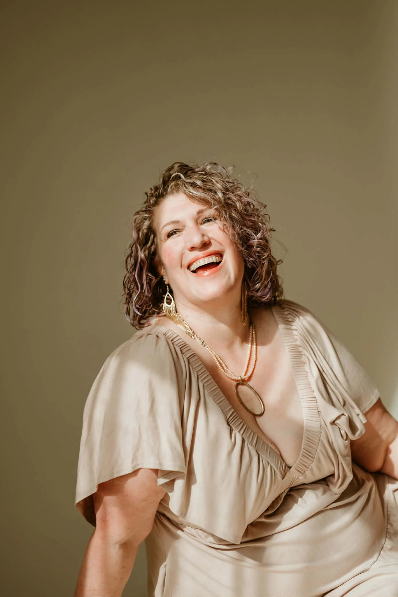 Page with curly hair, wearing a mauve dress and jewelry, is smiling and laughing against a plain background.