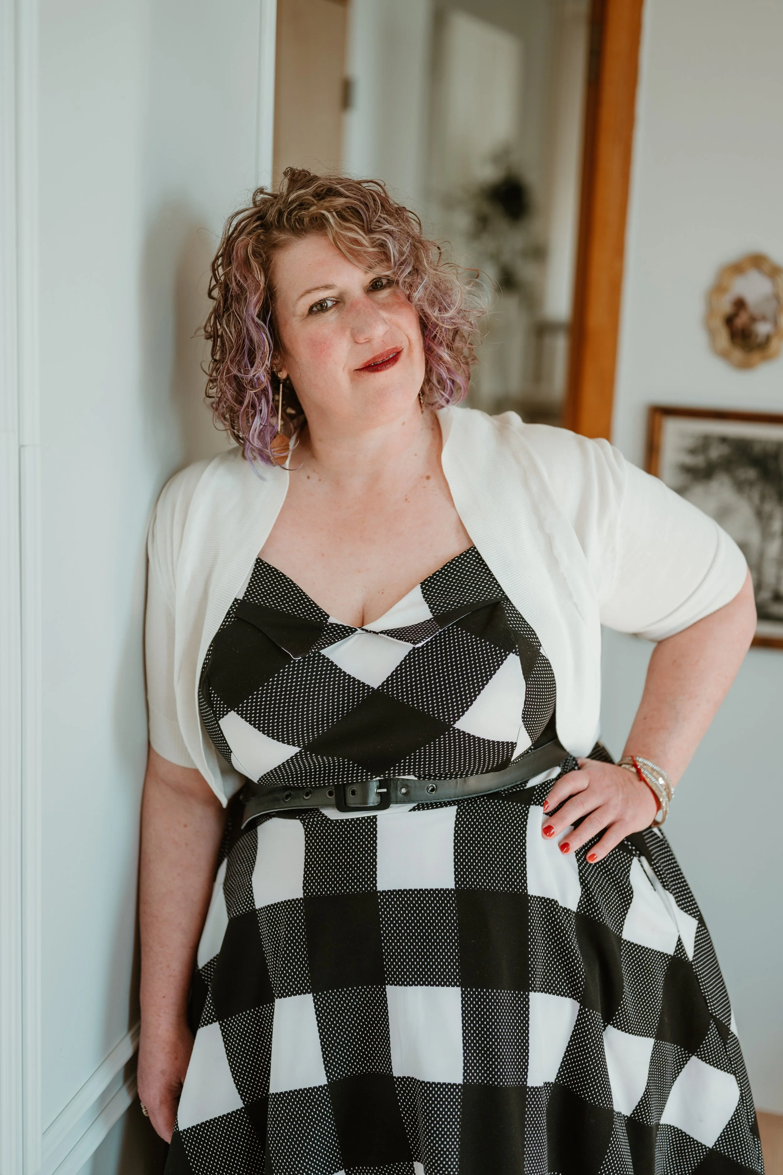 A woman with curly, purple-tinted hair in a black and white checkered dress, standing indoors with a confident expression.