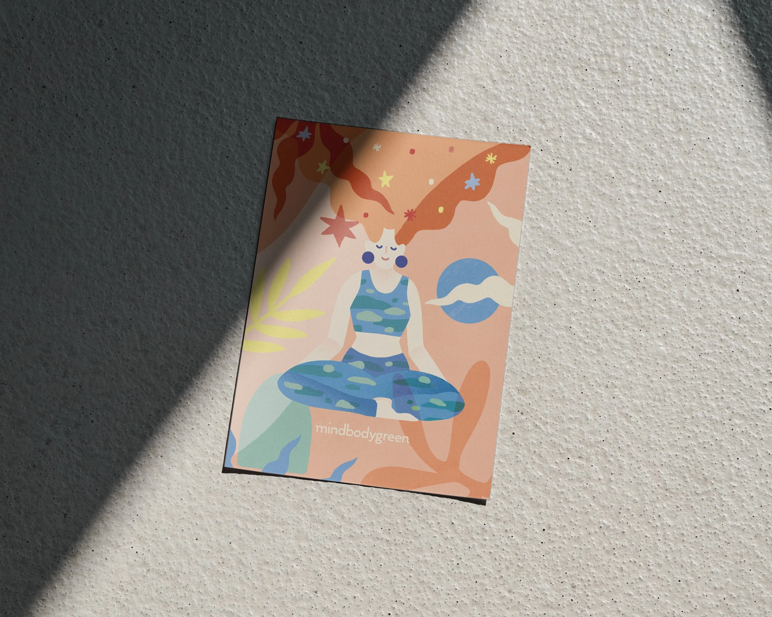 Colorful illustrated card featuring a woman with closed eyes, wearing a striped top, with floating stars and celestial elements in the background. The card displays the text "mindbodygreen" at the bottom.