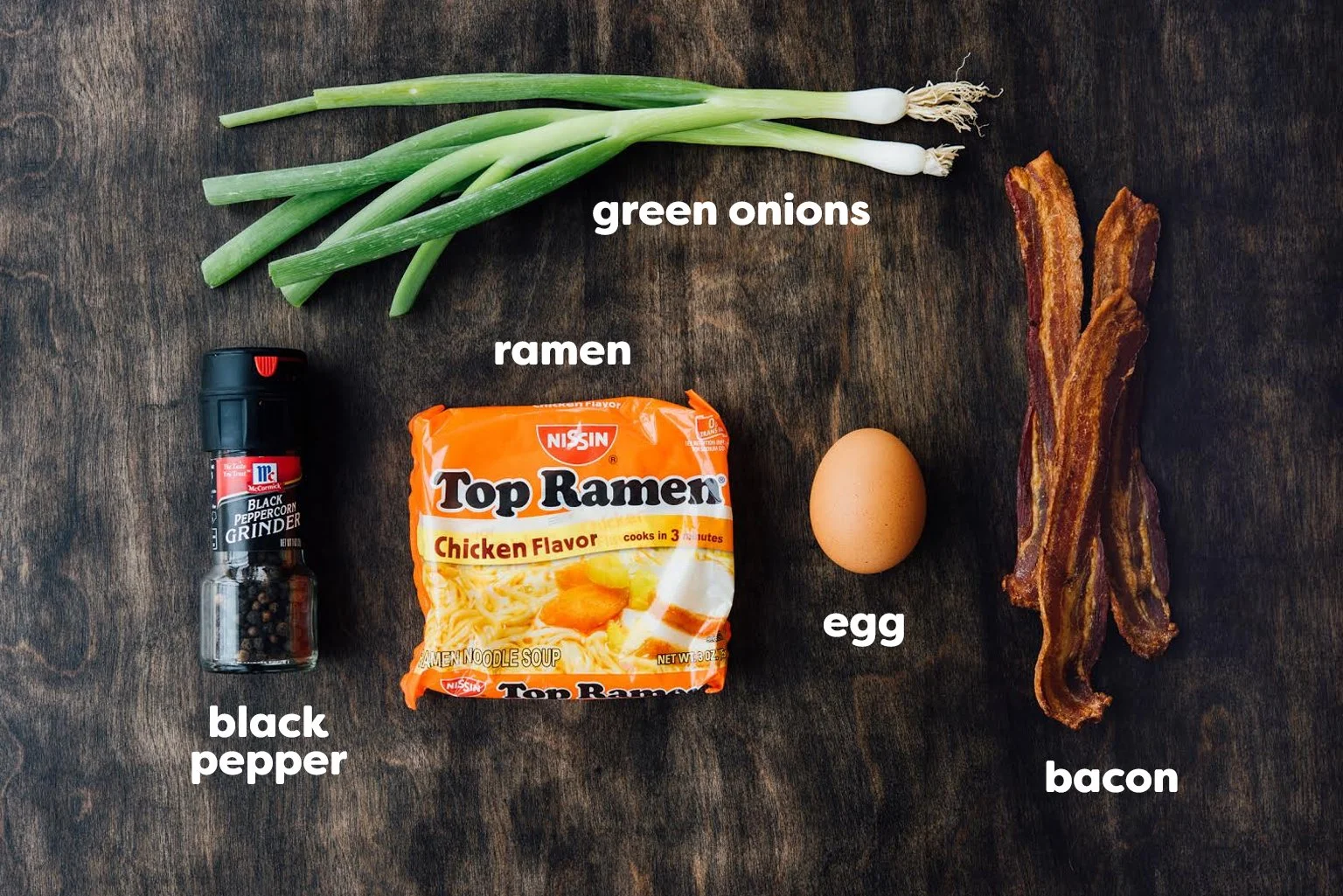 Green onions, ramen, egg, bacon, black pepper container on a wooden surface.
