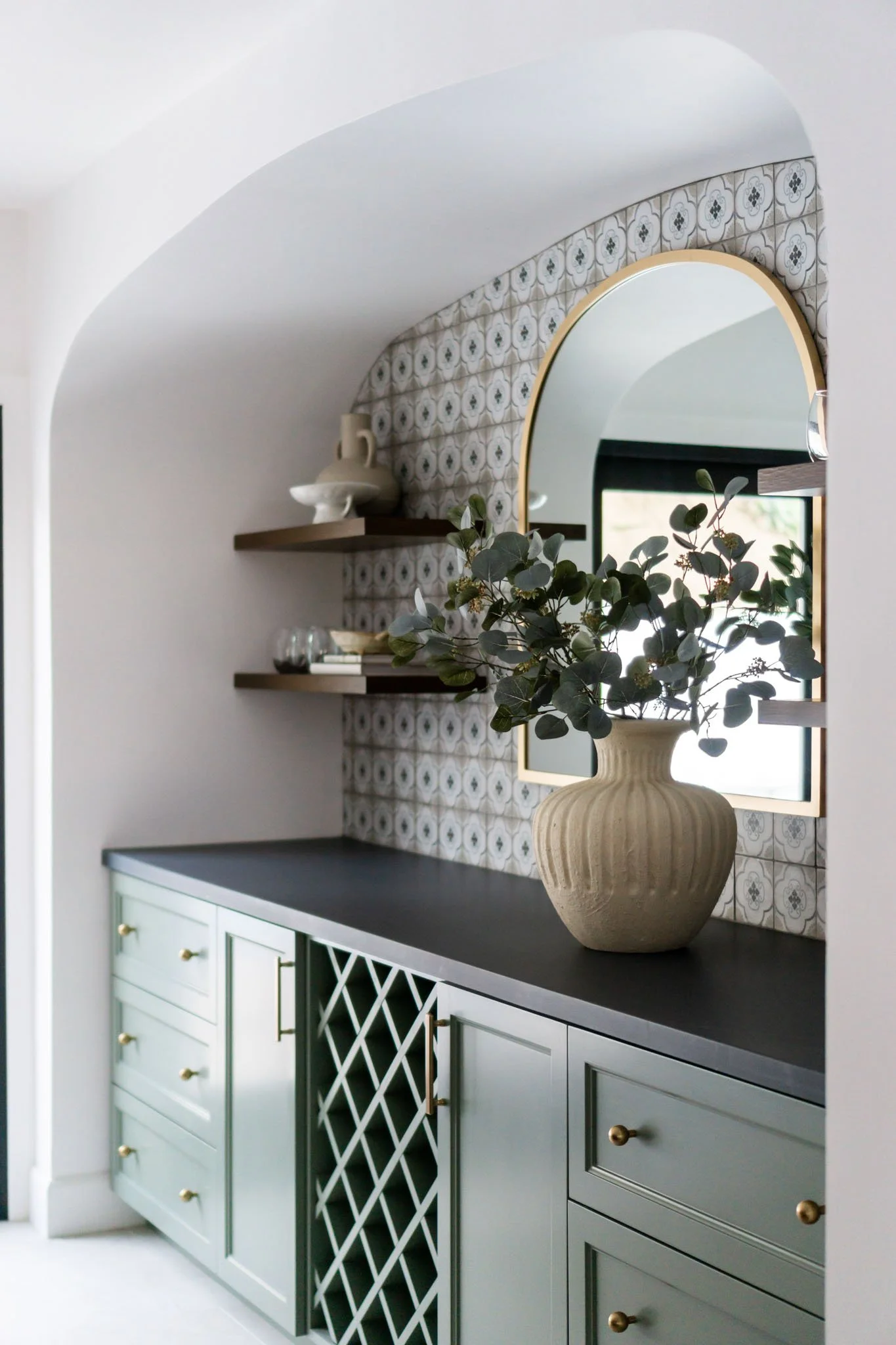 Stylish kitchen interior featuring green cabinets with brass handles, a dark countertop, a wine rack, and a tiled backsplash. A decorative vase with greenery is on the counter, and there are wooden shelves with ceramics and glassware. A round mirror is also featured.
