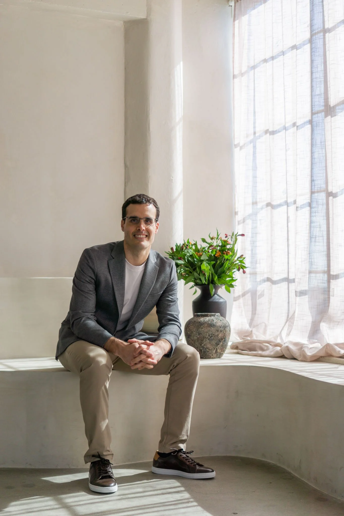 Man sitting on a white bench with a potted plant, wearing glasses, a gray blazer, white shirt, khaki pants, and dark shoes in a well-lit room with sheer curtains.