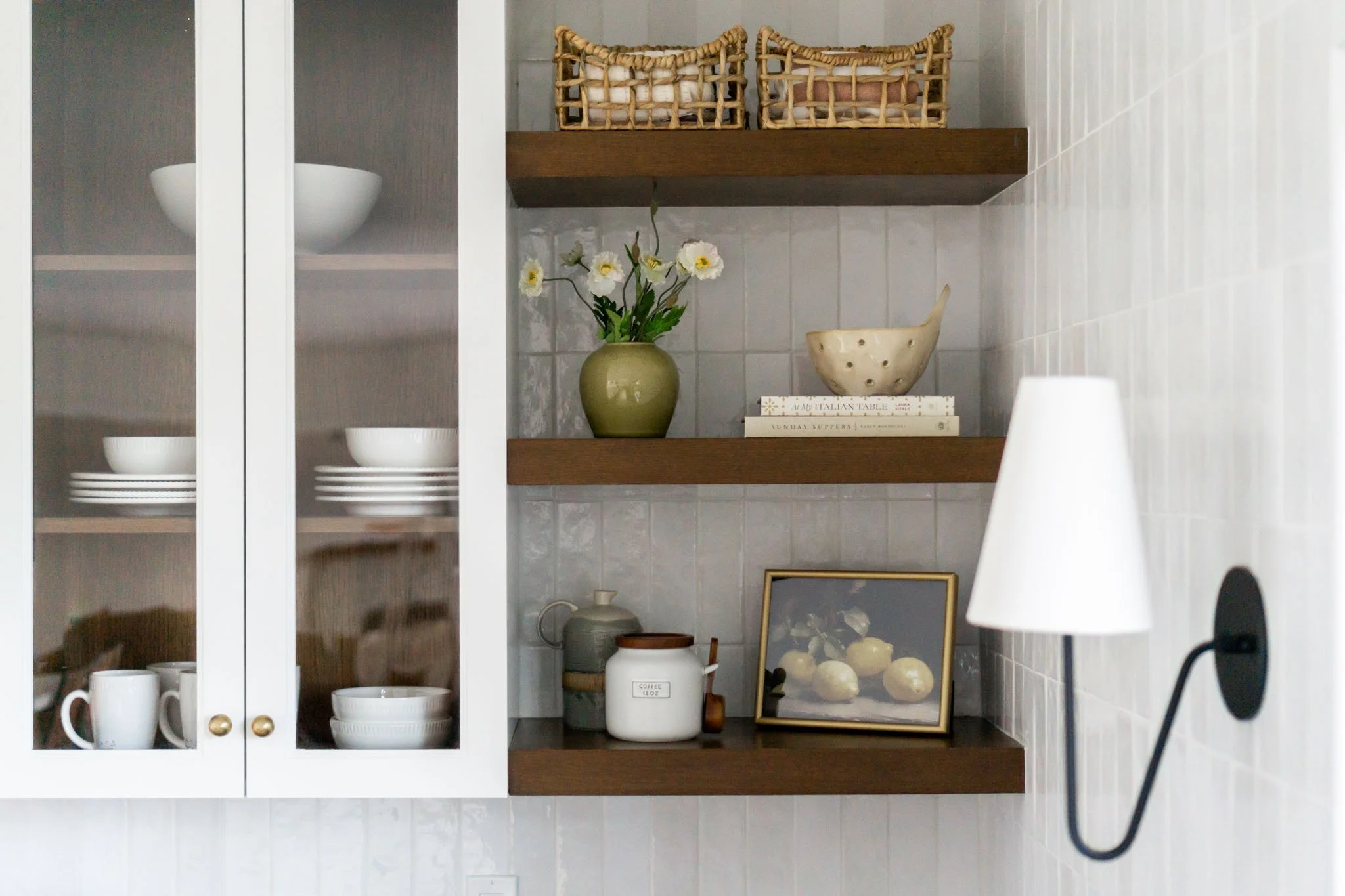 Kitchen shelves with dishes, wicker baskets, books, a green vase with flowers, a decorative bowl, and a framed picture; adjacent is a wall lamp.