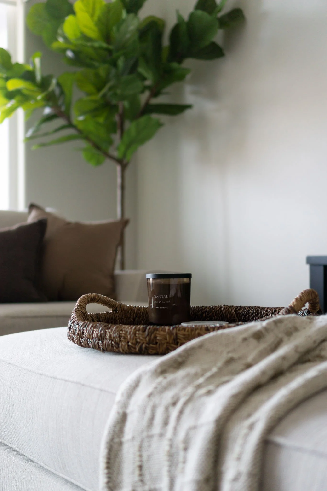 Cozy living room setup with a woven tray holding a candle on a beige ottoman, a soft knitted blanket draped over the side, and a green plant in the background.