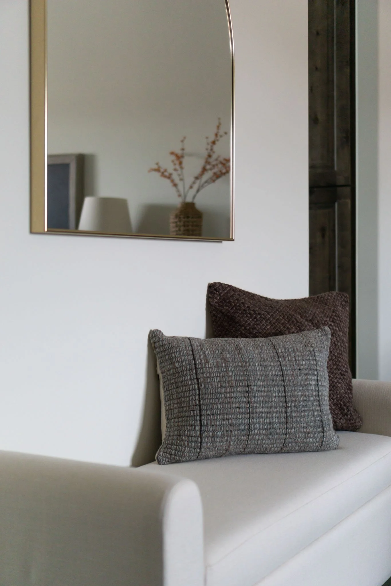 Neutral-toned room with a beige couch, two textured throw pillows, a wall mirror reflecting a vase with branches, and soft lighting.