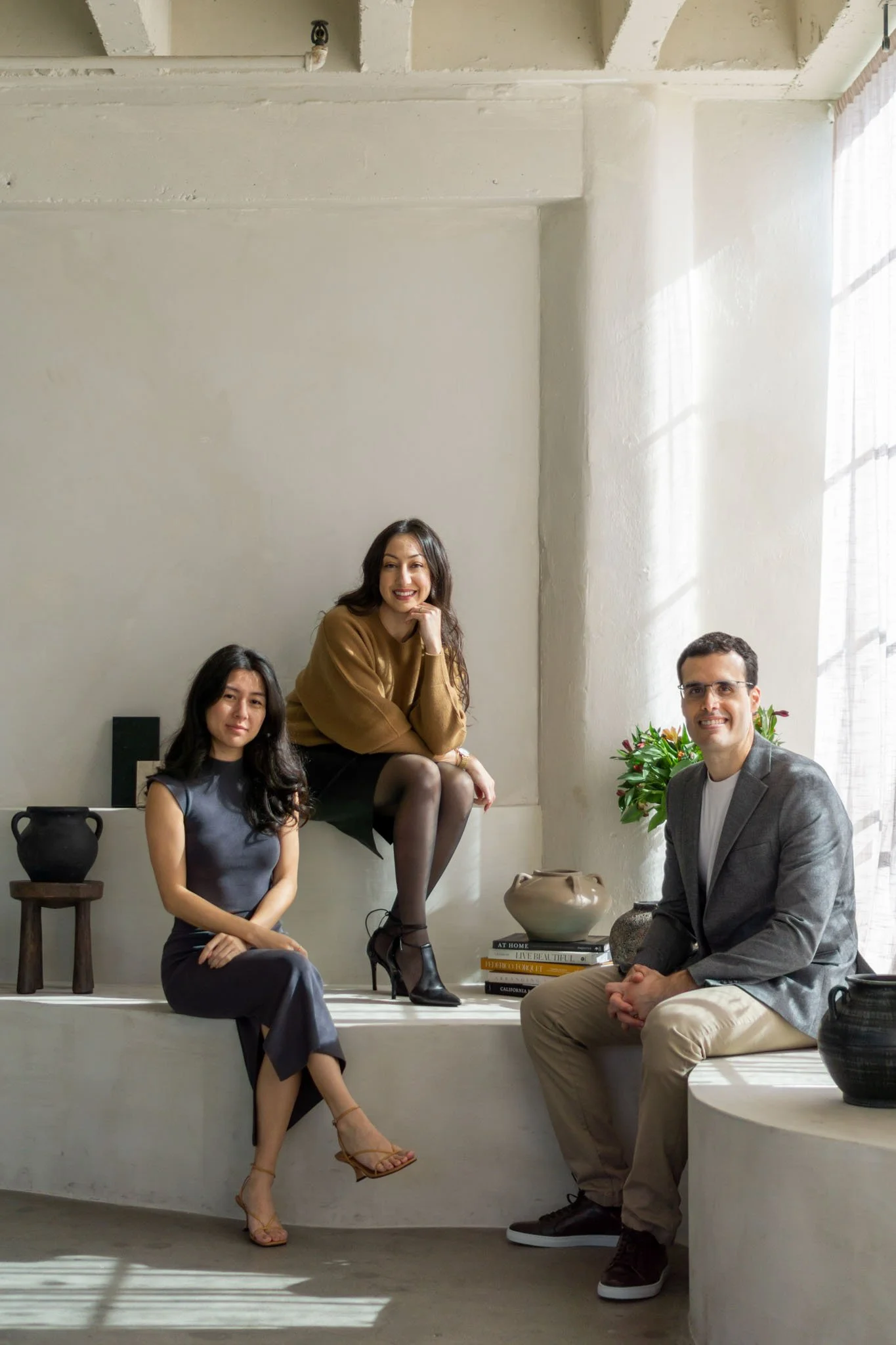 Three people sitting in a minimalist room with neutral decor. A woman in a blue dress sits on a ledge, another woman in a brown sweater and black skirt sits next to her, and a man in a gray blazer and beige pants is seated on a step. Decorative items and books are in the background.