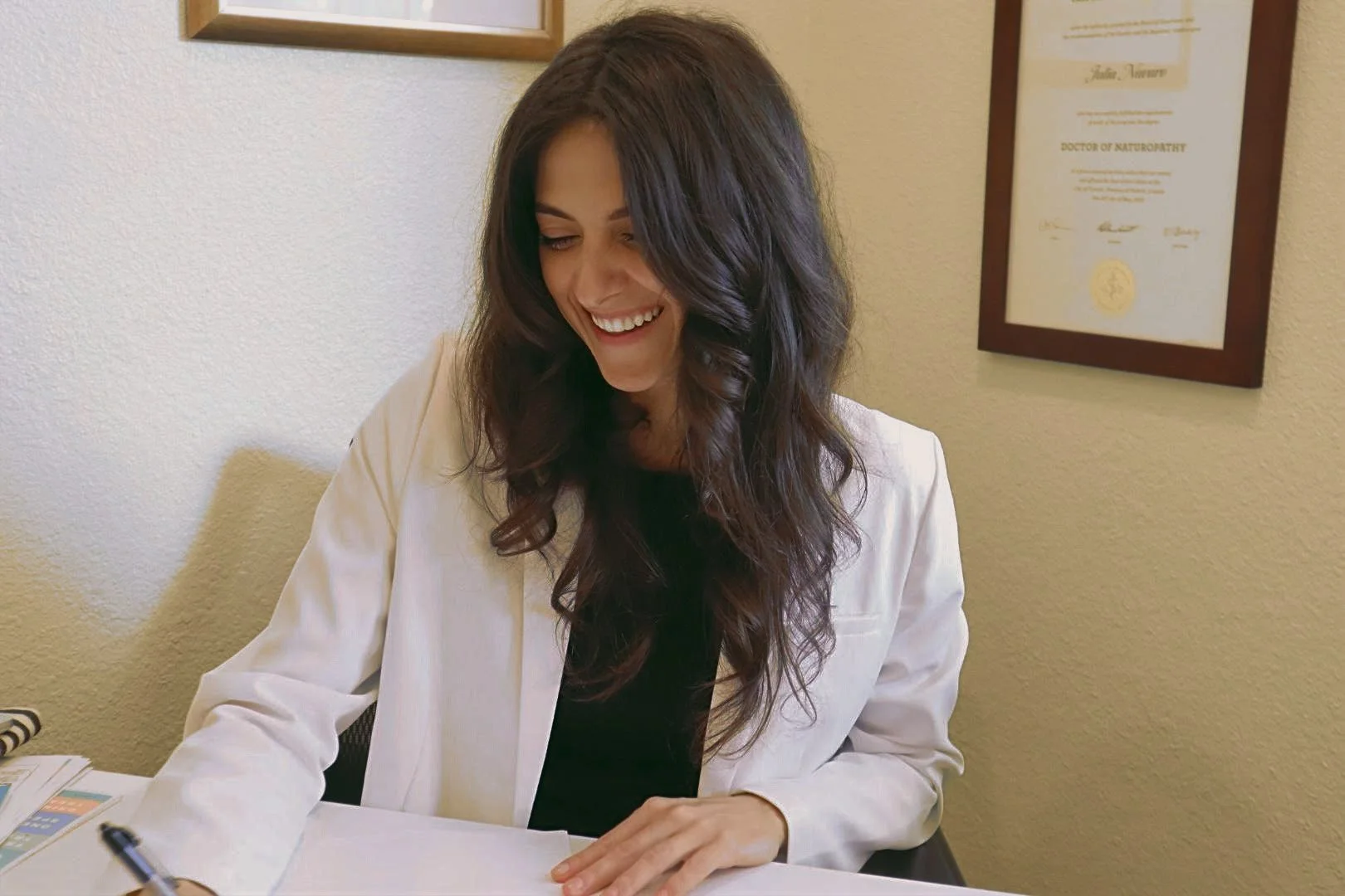 Smiling woman in white coat writing at a desk with framed certificates on the wall.