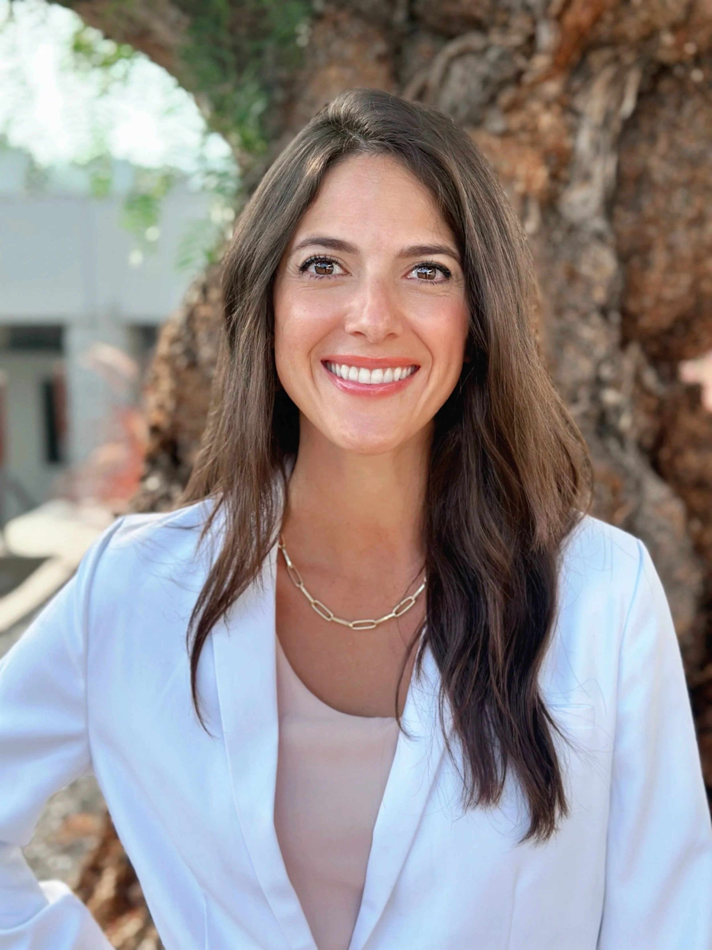 Smiling woman in a white blazer with long brown hair standing outdoors in front of a tree.