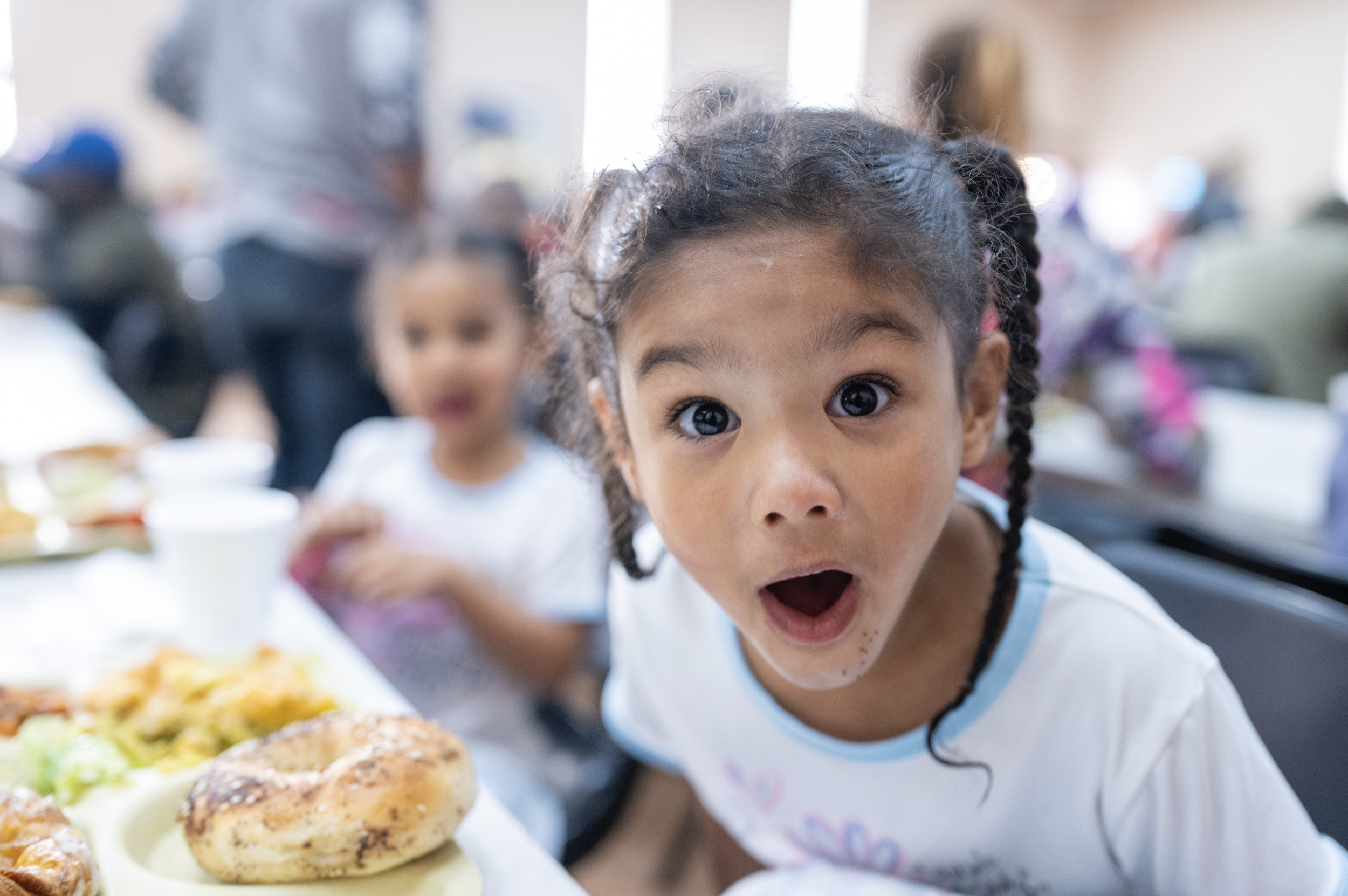 A young girl with braids looking surprised at the camera at a table with food, with another child in the background at a busy gathering or restaurant.