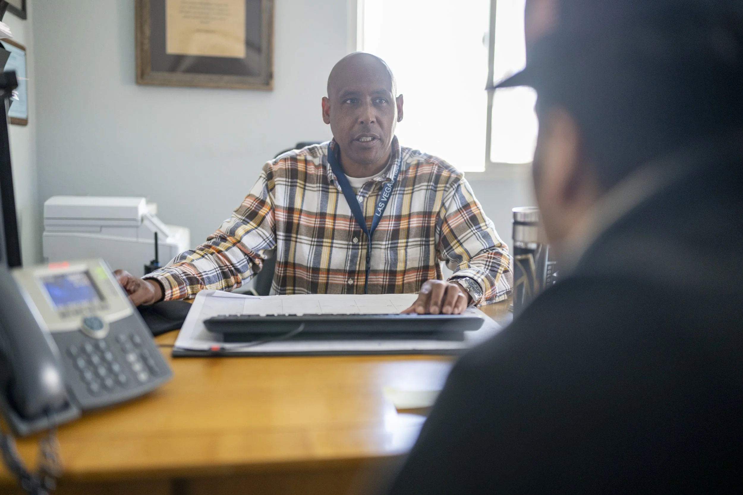 Two men having a conversation in an office, one man with a shaved head wearing a plaid shirt, another man with glasses seen from behind. There is a phone, a keyboard, and a printer on the desk.