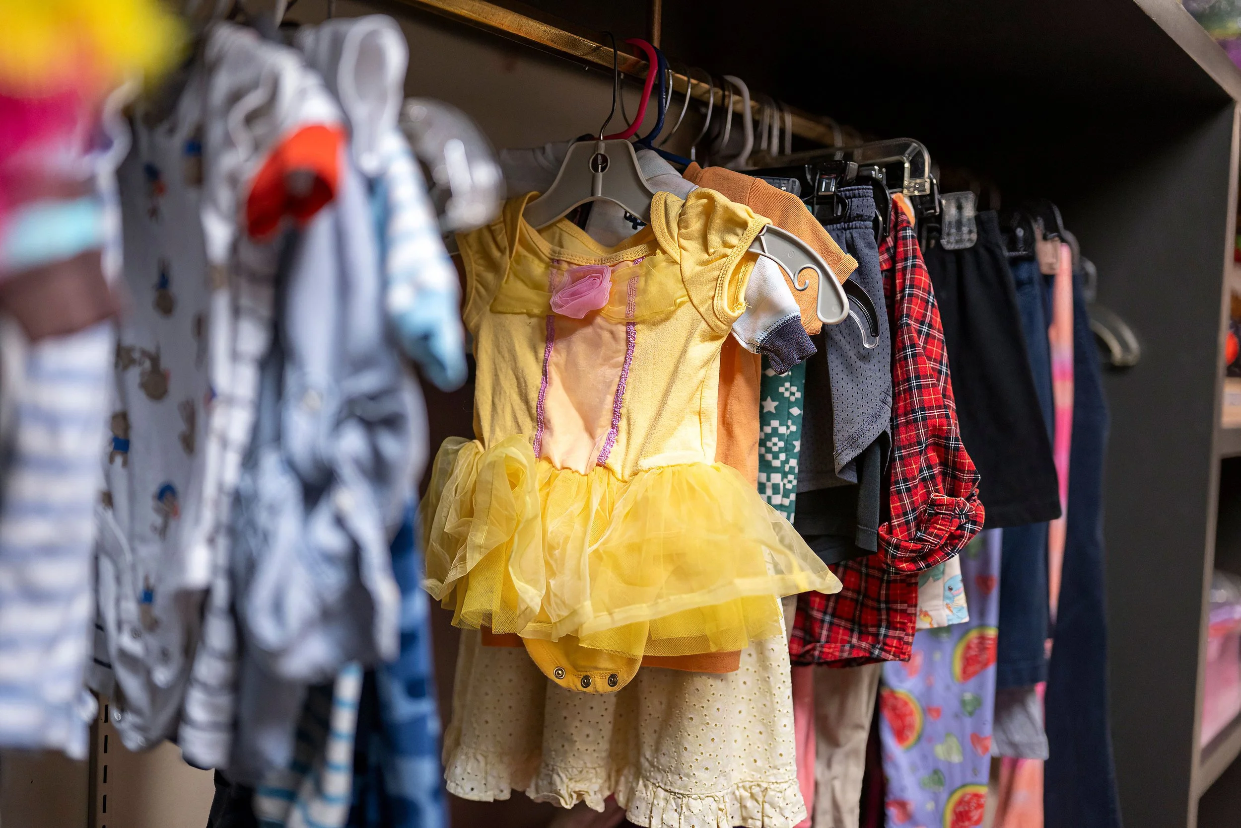 A row of children's clothes hanging on a rack, including a yellow princess dress, plaid shirts, and various patterned outfits.