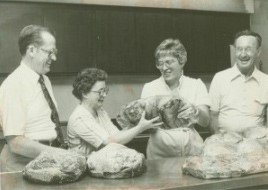 Four people, three men and one woman, standing at a table, smiling and holding packages of food in a kitchen or dining area.