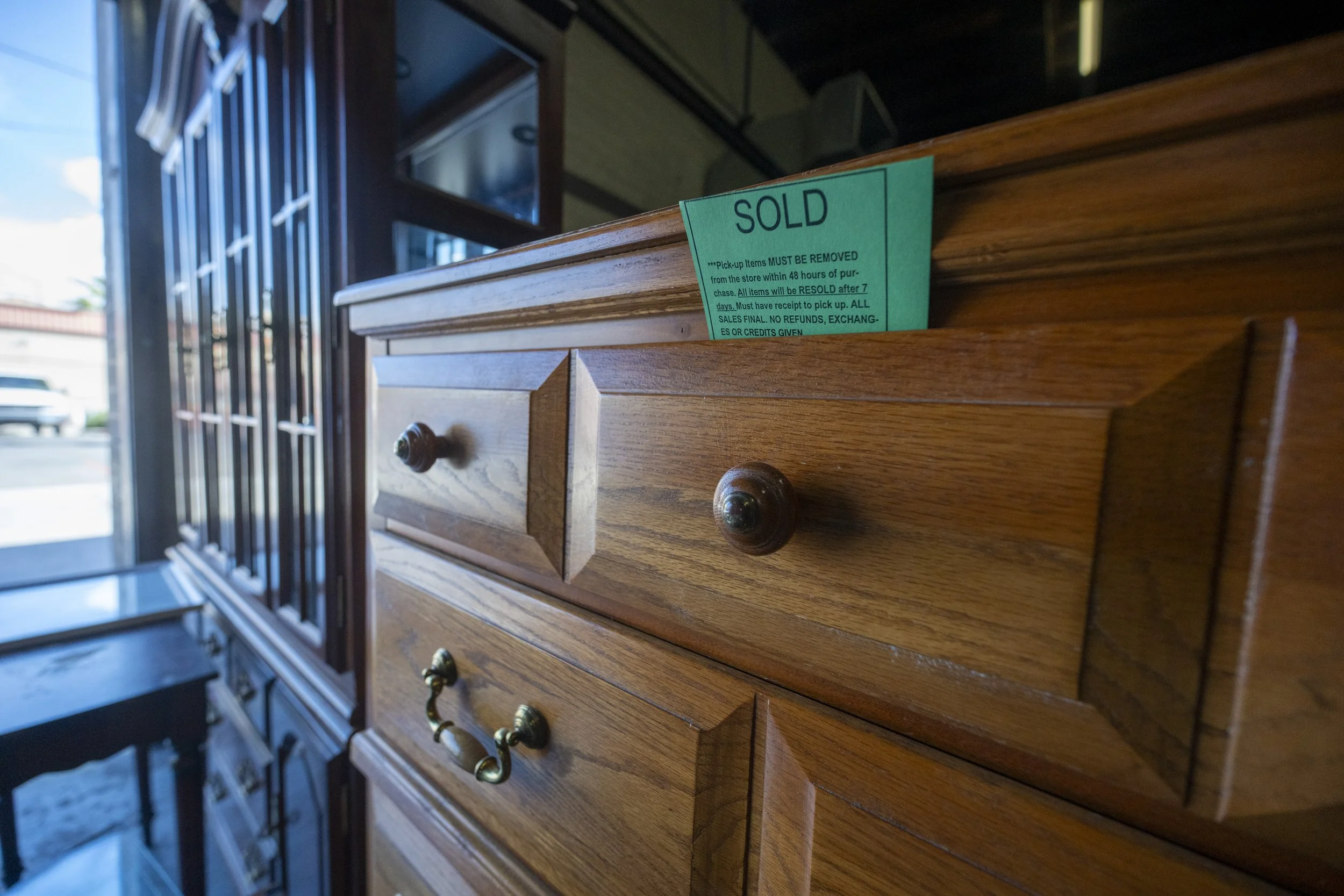 Wooden furniture with a green 'SOLD' tag on top, inside a store with a glass door and window.