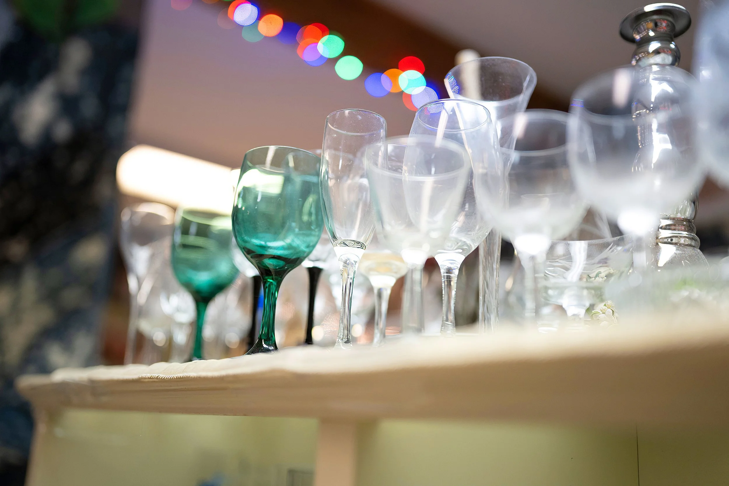 A collection of empty and partially filled wine glasses and decanter on a wooden shelf, with colorful blurred string lights in the background.