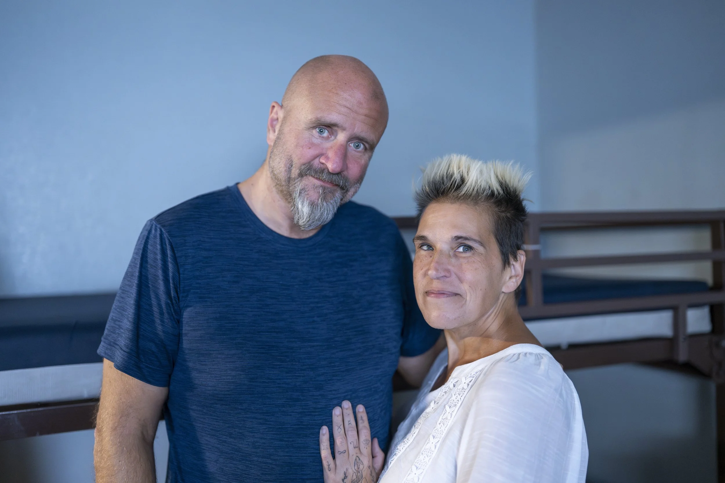 A middle-aged man with a beard and a woman with short, styled hair standing close together indoors, with their hands touching.