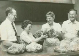 Four people standing behind a table with bags, smiling and handing a loaf of bread to a woman in the middle, in a vintage black-and-white setting.
