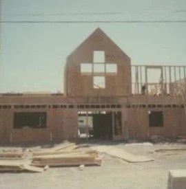 A brick building under construction with a large white cross symbol on the upper front wall, and construction materials in front.