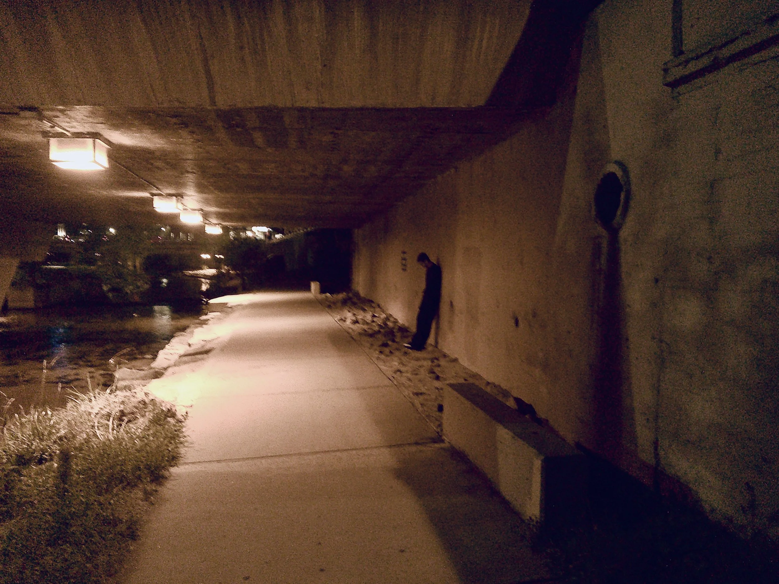 Dimly lit nighttime walkway beside a building, with glowing wall lamps, snowy ground, and a silhouetted person.