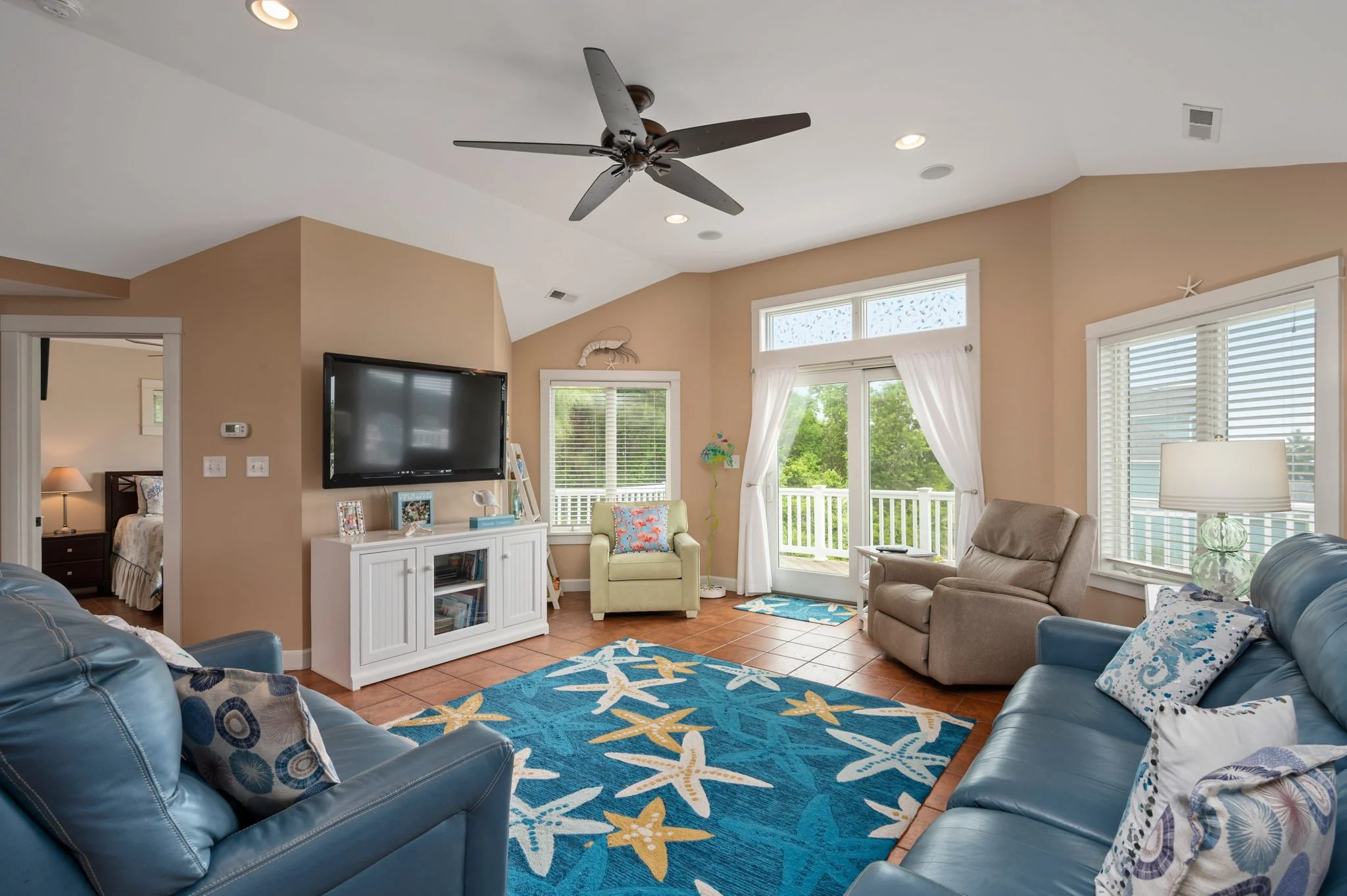 Coastal-themed living room with blue leather sofa, patterned pillows, TV on beige wall, ceiling fan, armchairs, and glass doors leading to balcony.