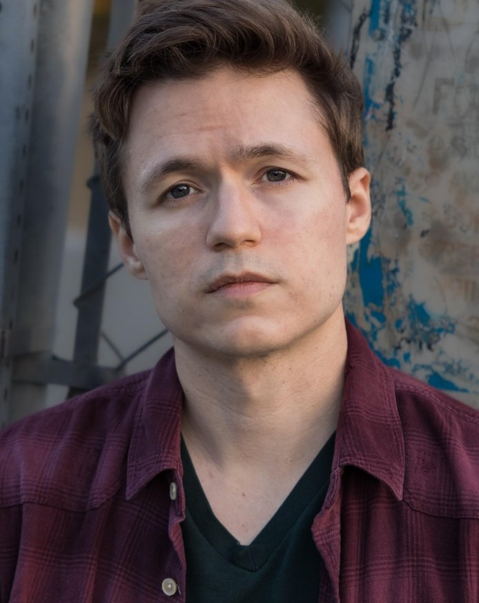 A young man with short brown hair and fair skin, wearing a maroon flannel over a green V-neck shirt, looking directly at the camera with a neutral expression, outdoors with a background of metal and painted surface.