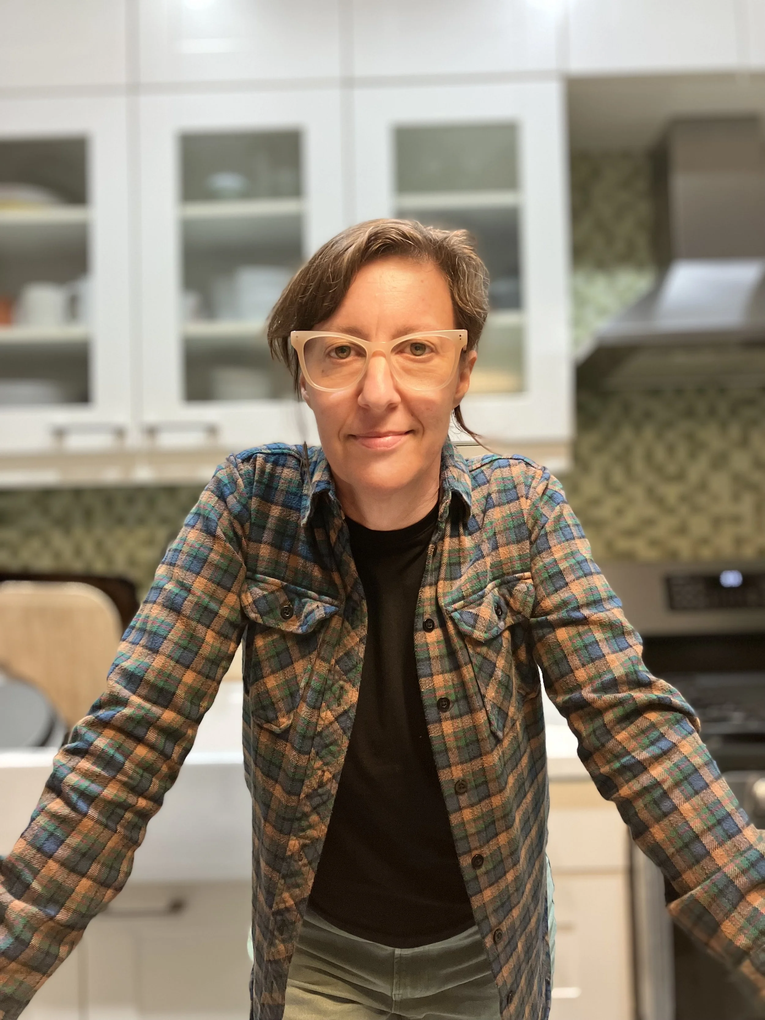 A woman with short brown hair, wearing beige glasses, a plaid shirt over a black top, standing in a kitchen with white cabinets, a stove, and a green patterned backsplash.