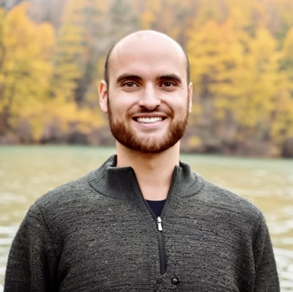 Smiling man outdoors near a body of water with trees in autumn colors in the background.