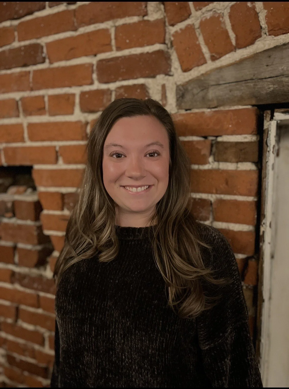A young woman with long wavy brown hair smiling, standing in front of a brick fireplace.