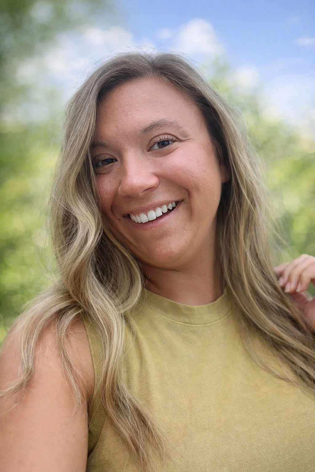 Close-up of a smiling woman outdoors with a blue sky and green trees in the background.