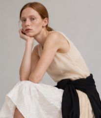 A young woman sitting with her head resting on her hand, wearing a sleeveless cream top and a white skirt, against a plain gray background.