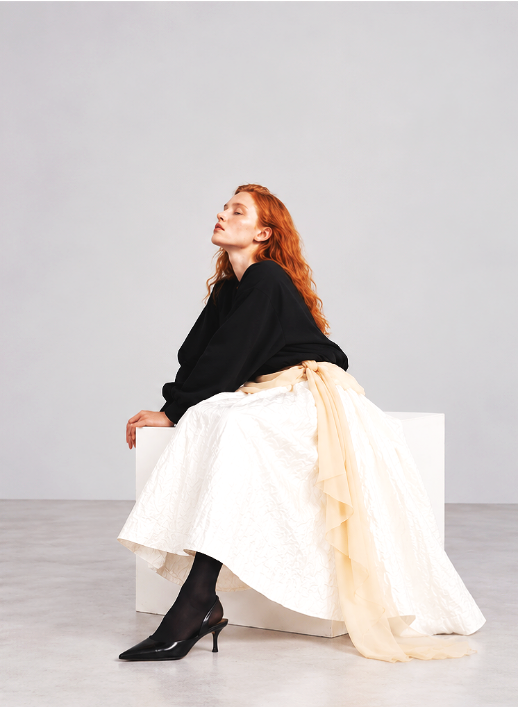 A woman with red hair wearing a black top, cream-colored skirt with a sash, black stockings, and high heels, sitting on a white block against a plain gray background.